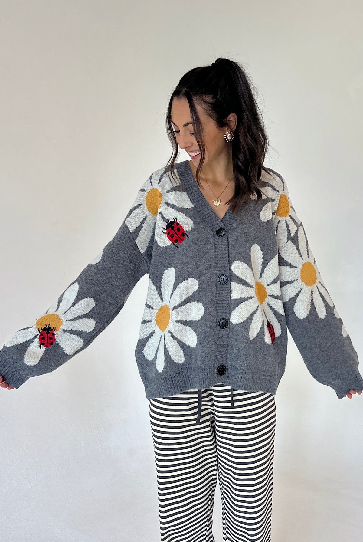 A woman modeling a gray knit cardigan with large white daisy patterns, yellow centers, and red ladybug motifs, paired with black and white striped pants against a plain light background.