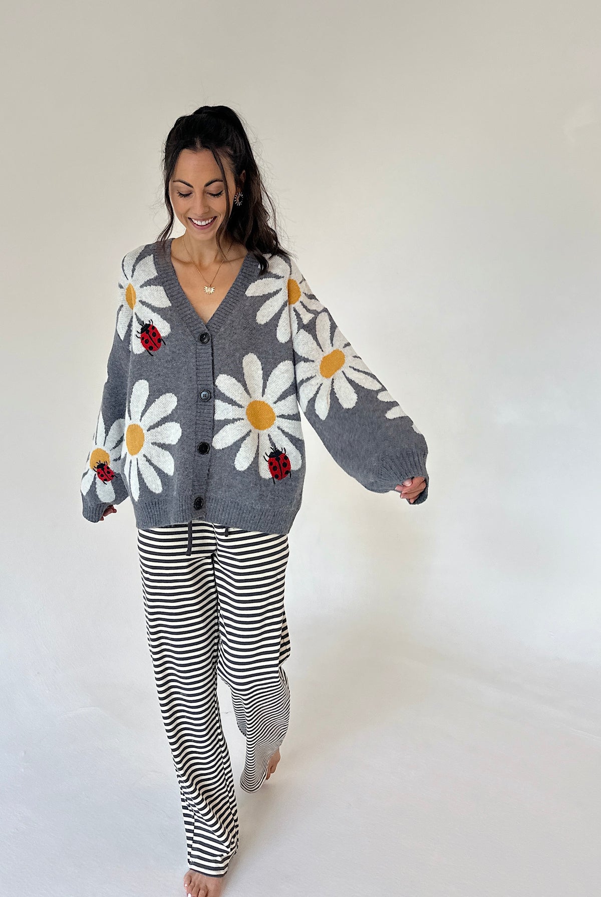 A woman modeling a cozy gray knit cardigan with large white daisy patterns, yellow centers, and small red ladybugs, paired with black and white striped pajama pants, against a plain white studio background.