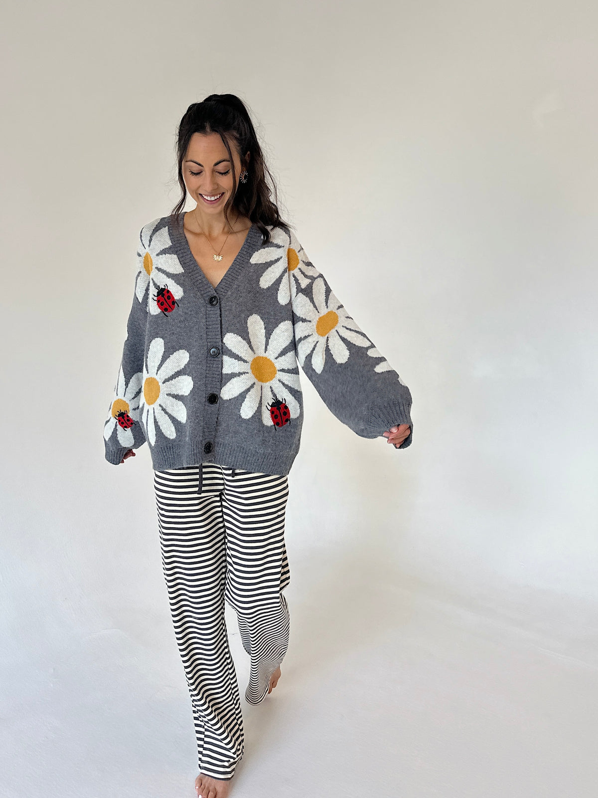 A woman modeling a cozy gray knit cardigan with large white daisy patterns, yellow centers, and small red ladybugs, paired with black and white striped pajama pants, against a plain white studio background.