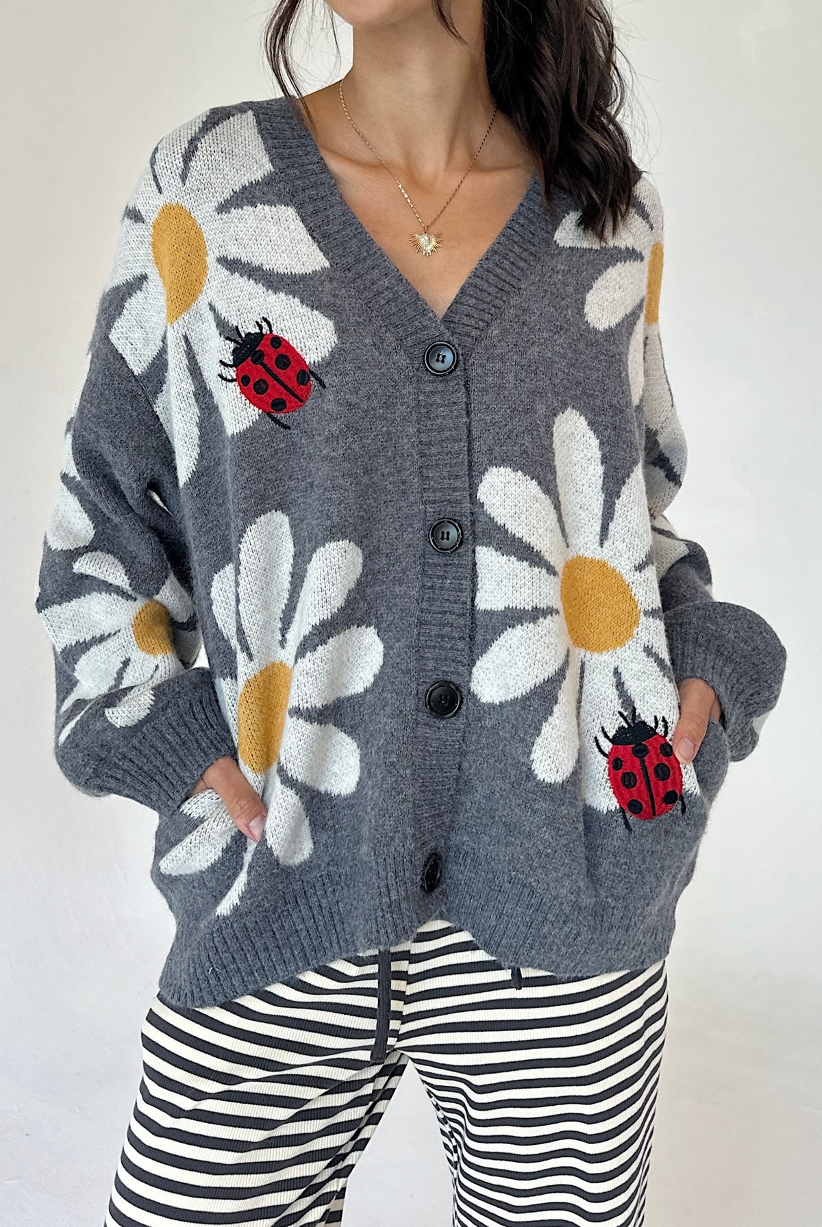 A woman wearing a gray knit cardigan with large white daisy patterns, yellow centers, and red ladybugs, paired with black and white striped pants against a plain light background.