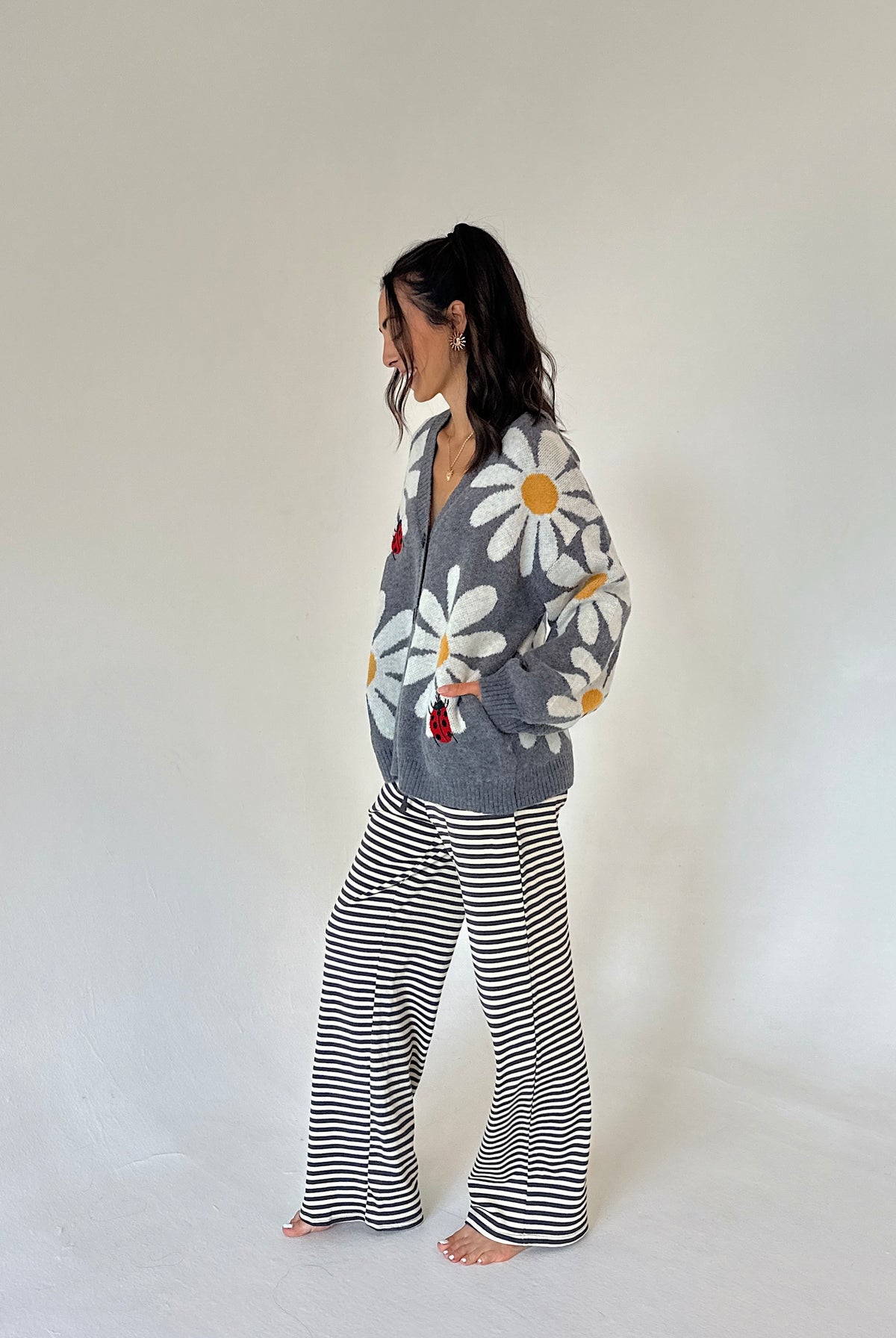 A woman stands barefoot in profile wearing a gray cardigan with a large white daisy print and black-and-white horizontally striped wide-leg pants against a plain off-white studio background.
