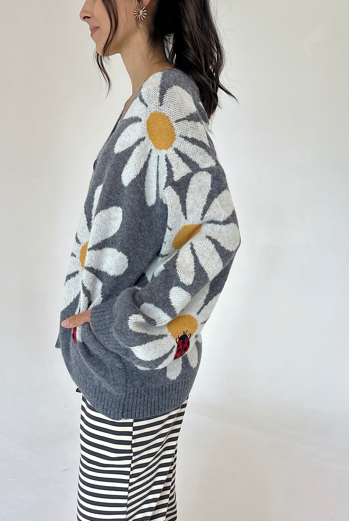 A woman wearing a gray knit cardigan with large white daisy patterns and yellow centers, paired with black and white striped pants, photographed against a plain white background.