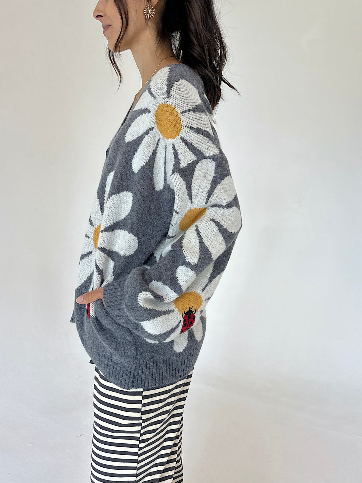 A woman wearing a gray knit cardigan with large white daisy patterns and yellow centers, paired with black and white striped pants, photographed against a plain white background.