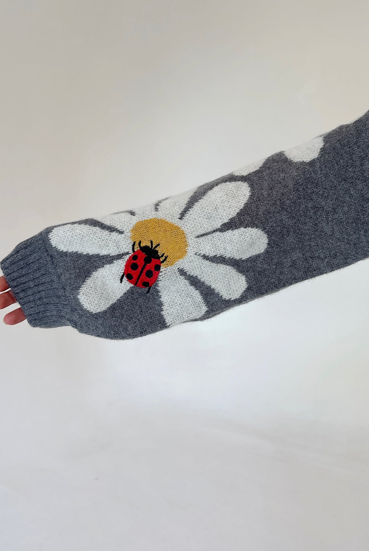 A close-up of a gray knit sleeve featuring a large white daisy with a yellow center and a red ladybug motif, held against a plain light background.