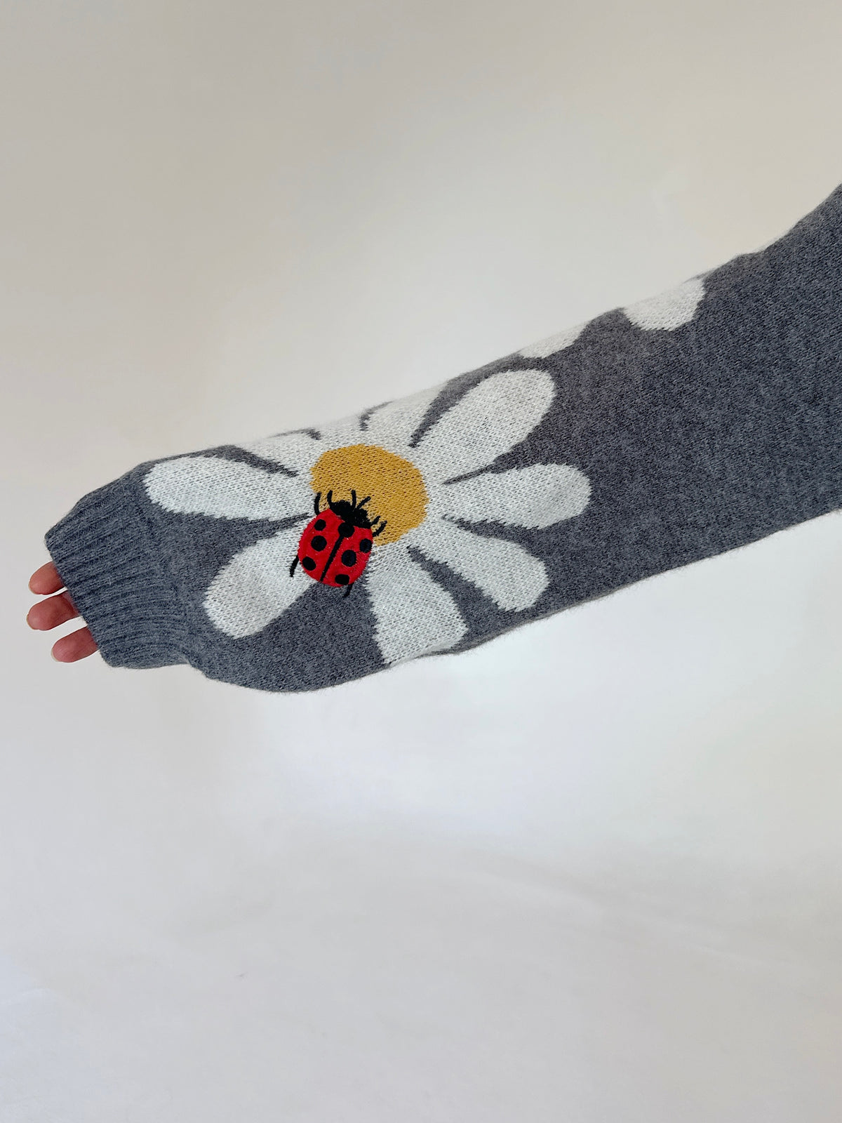 A close-up of a gray knit sleeve featuring a large white daisy with a yellow center and a red ladybug motif, held against a plain light background.