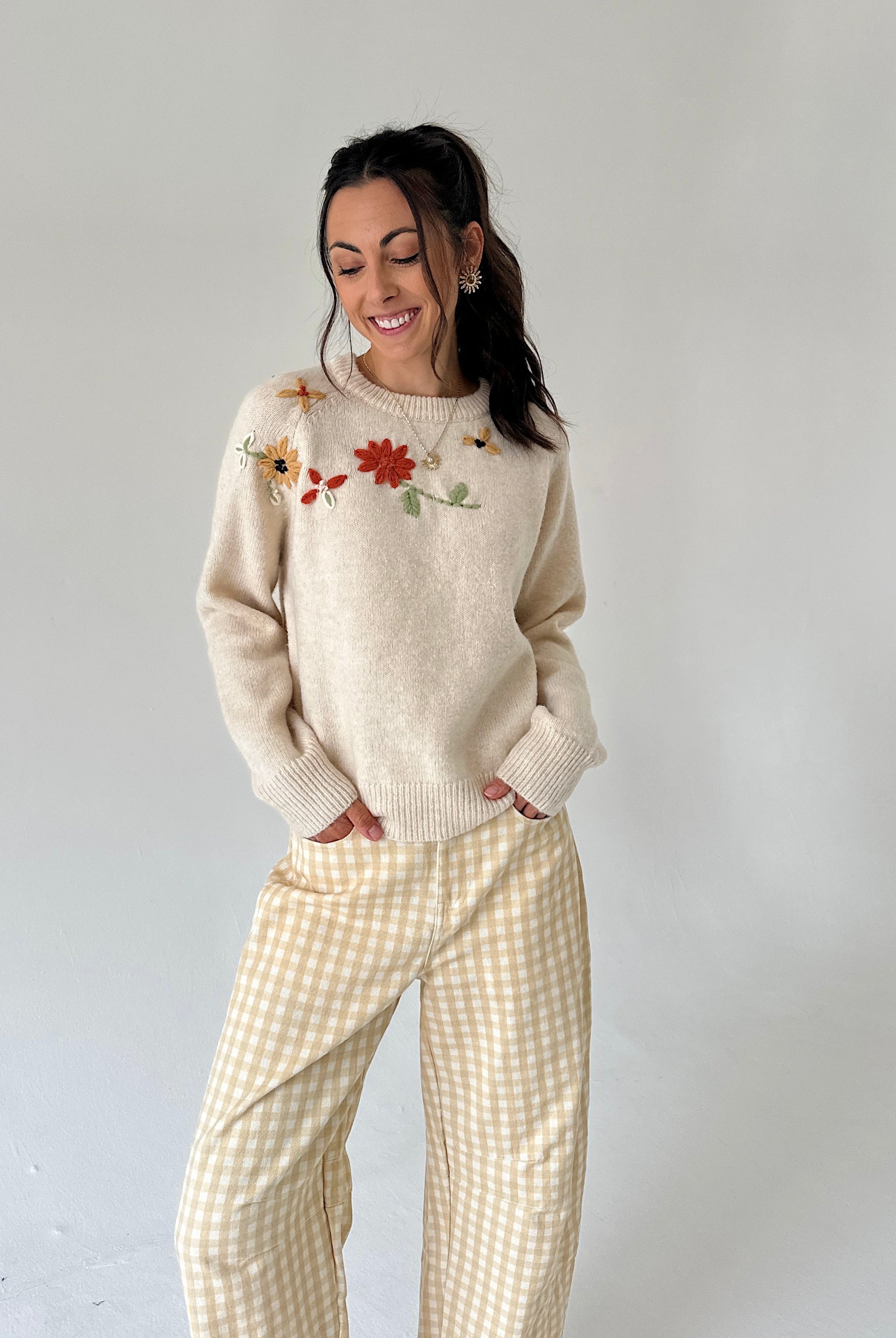 A woman modeling a cream-colored knit sweater with colorful floral embroidery on the chest, paired with yellow and white gingham wide-leg pants, against a plain light gray studio background.