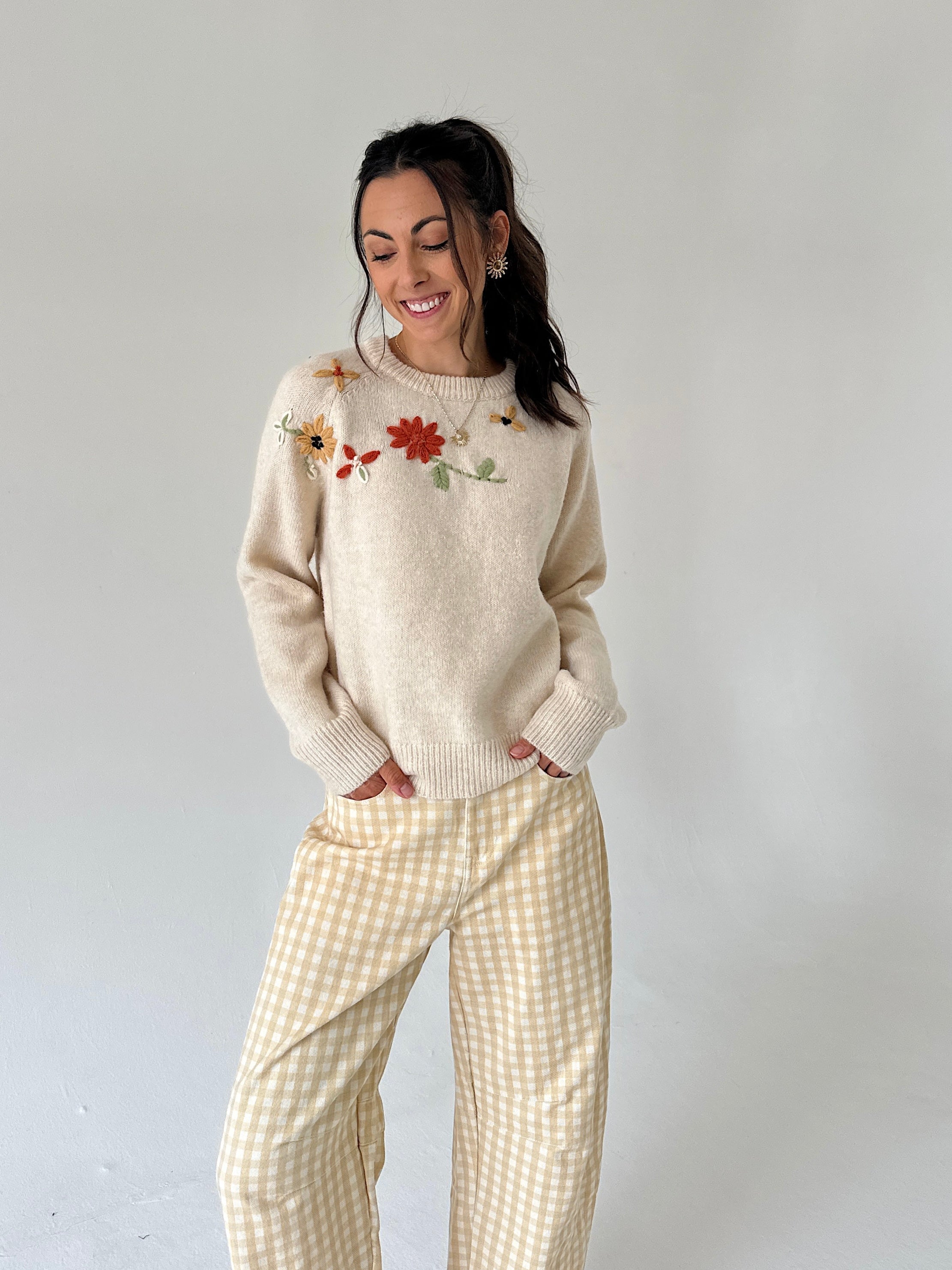 A woman modeling a cream-colored knit sweater with colorful floral embroidery on the chest, paired with yellow and white gingham wide-leg pants, against a plain light gray studio background.