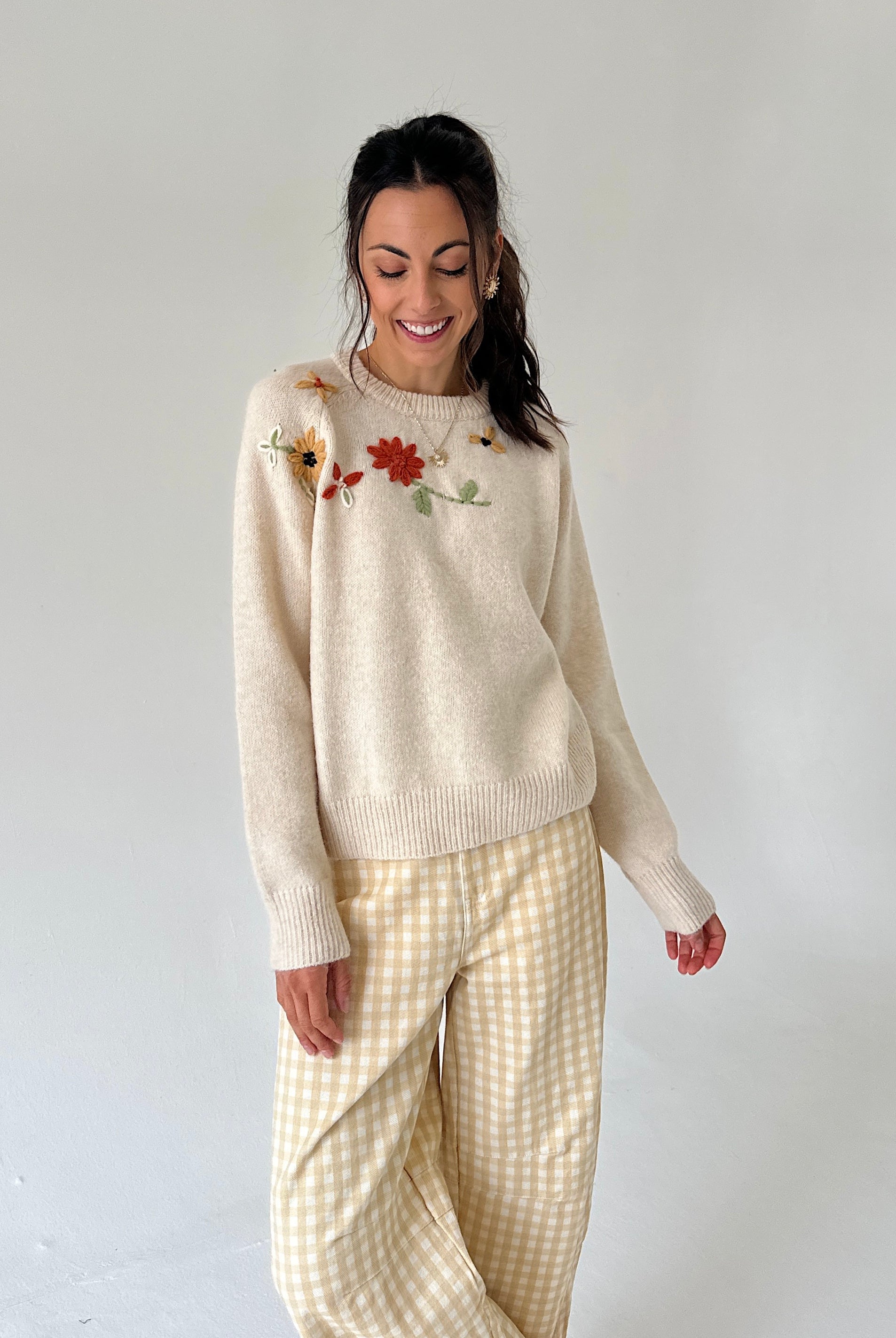 A woman modeling a cream-colored knit sweater with colorful floral embroidery on the chest, paired with cream and beige gingham pants, against a plain light gray background.