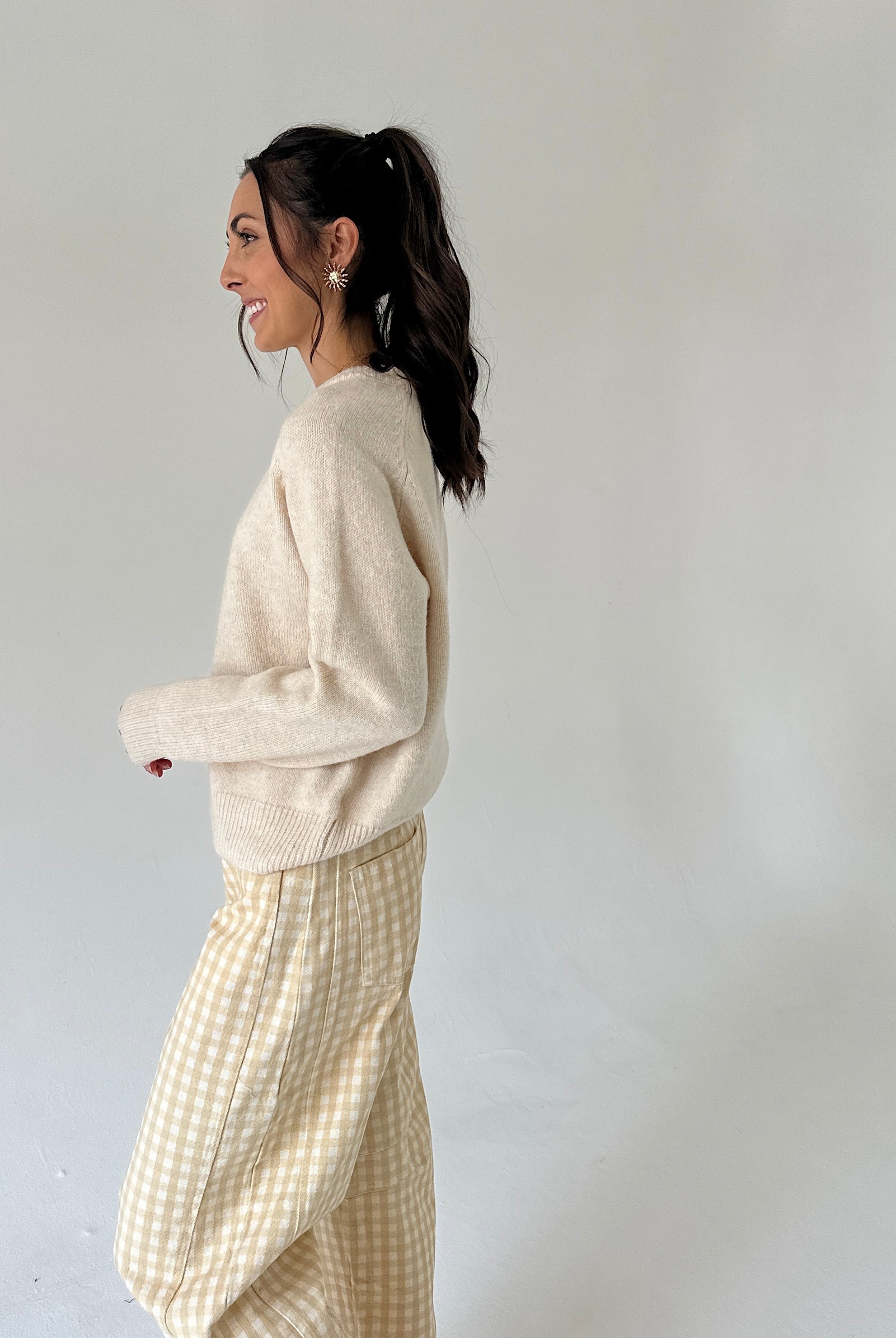 A woman in a side profile wearing a cream knit sweater and a beige and white gingham skirt, standing against a plain light gray studio background.
