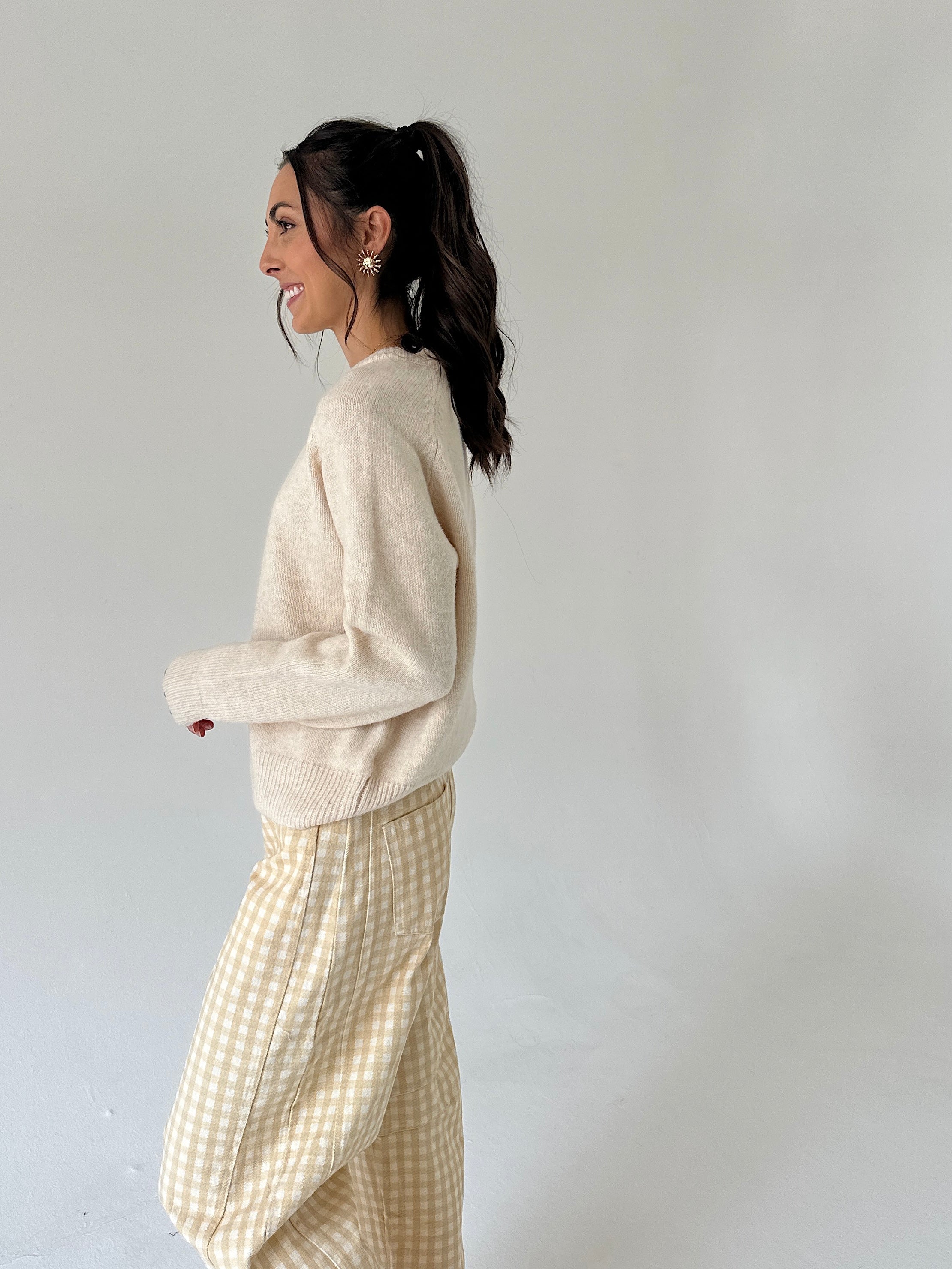 A woman in a side profile wearing a cream knit sweater and a beige and white gingham skirt, standing against a plain light gray studio background.