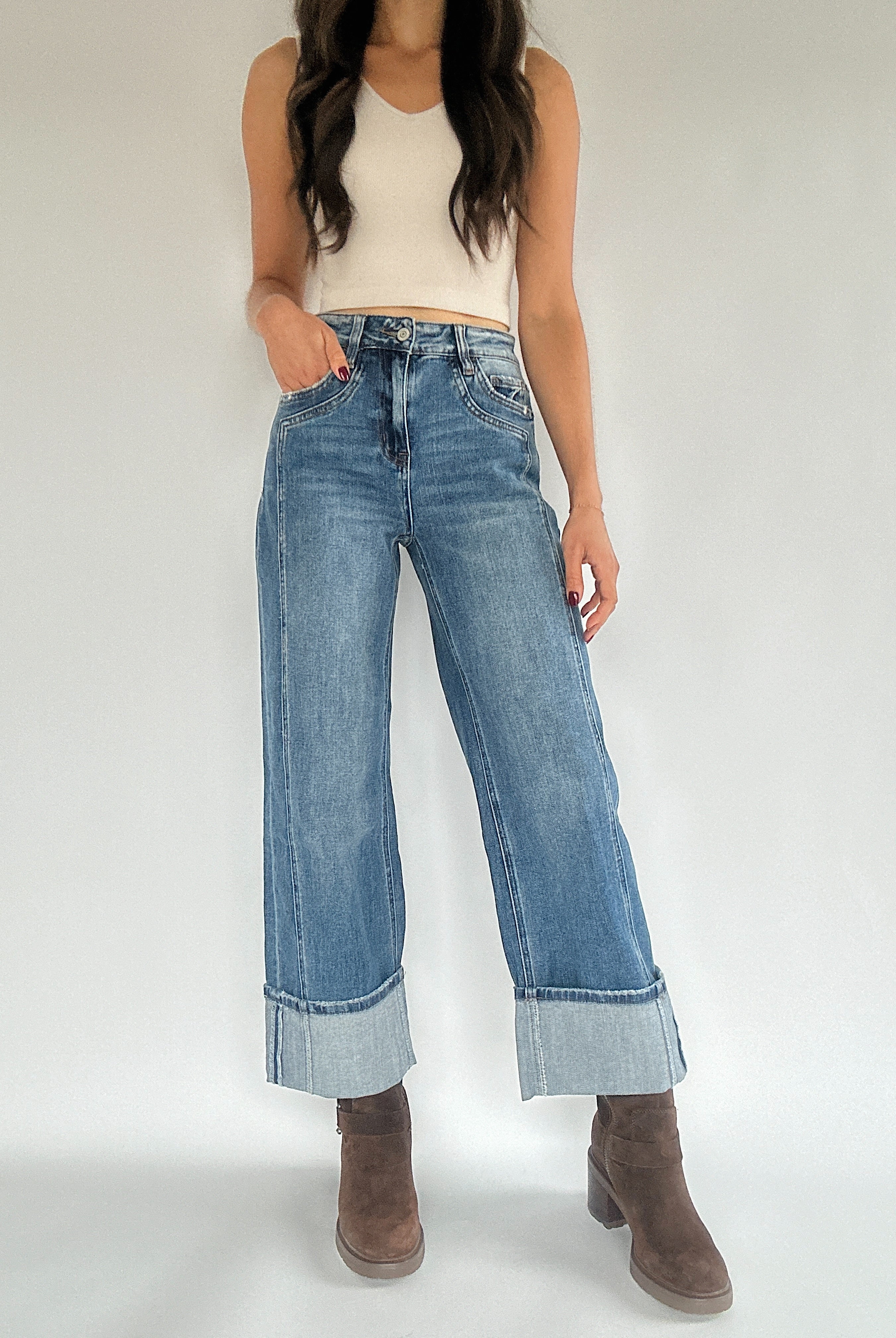 A person wearing medium washed denim jeans with a wide leg silhouette and a cuffed bottom, paired with a white sleeveless top and brown ankle boots.