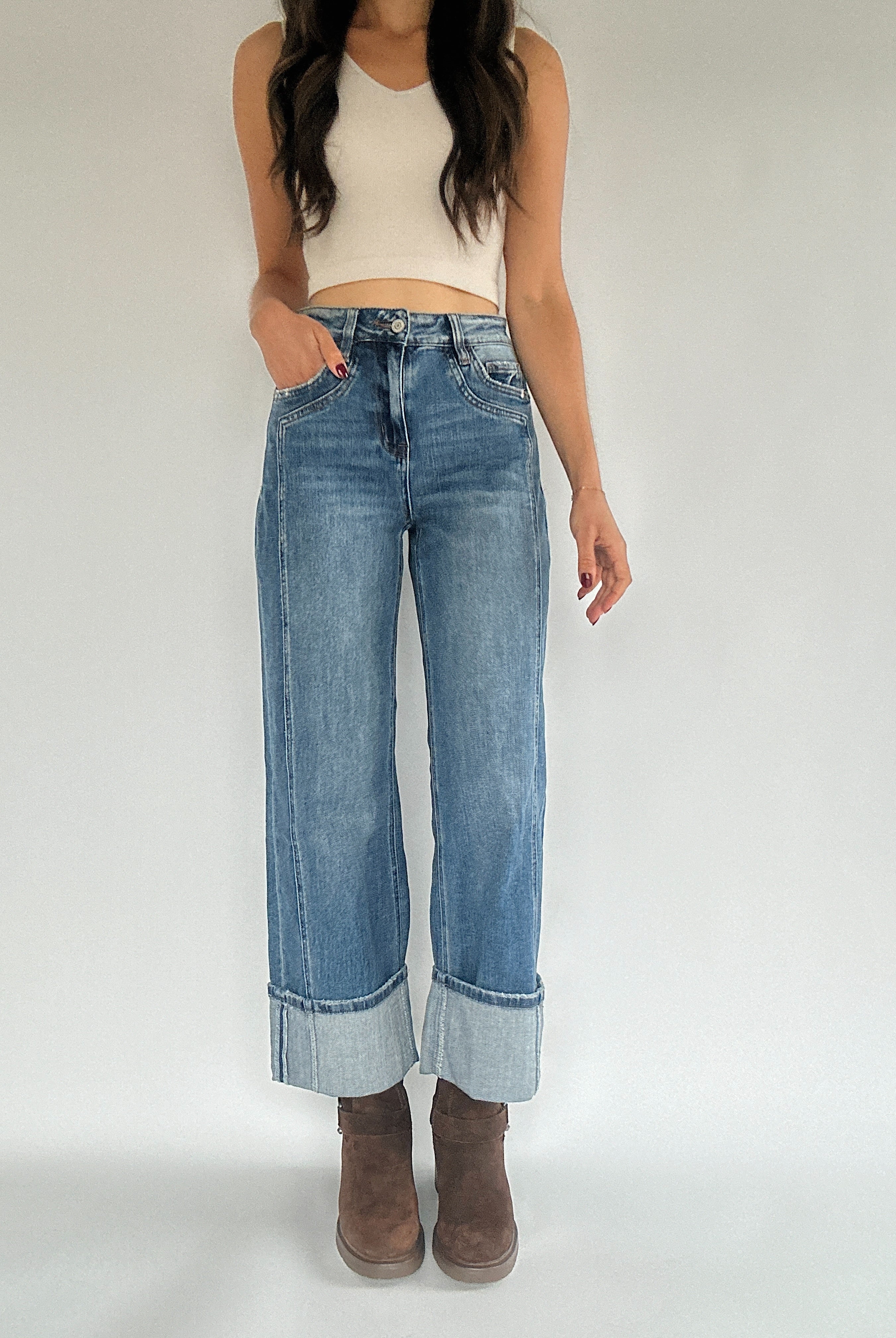 Woman wearing medium wash denim wide leg jeans that are cuffed at the bottom and paired with brown ankle books and a cream tank.