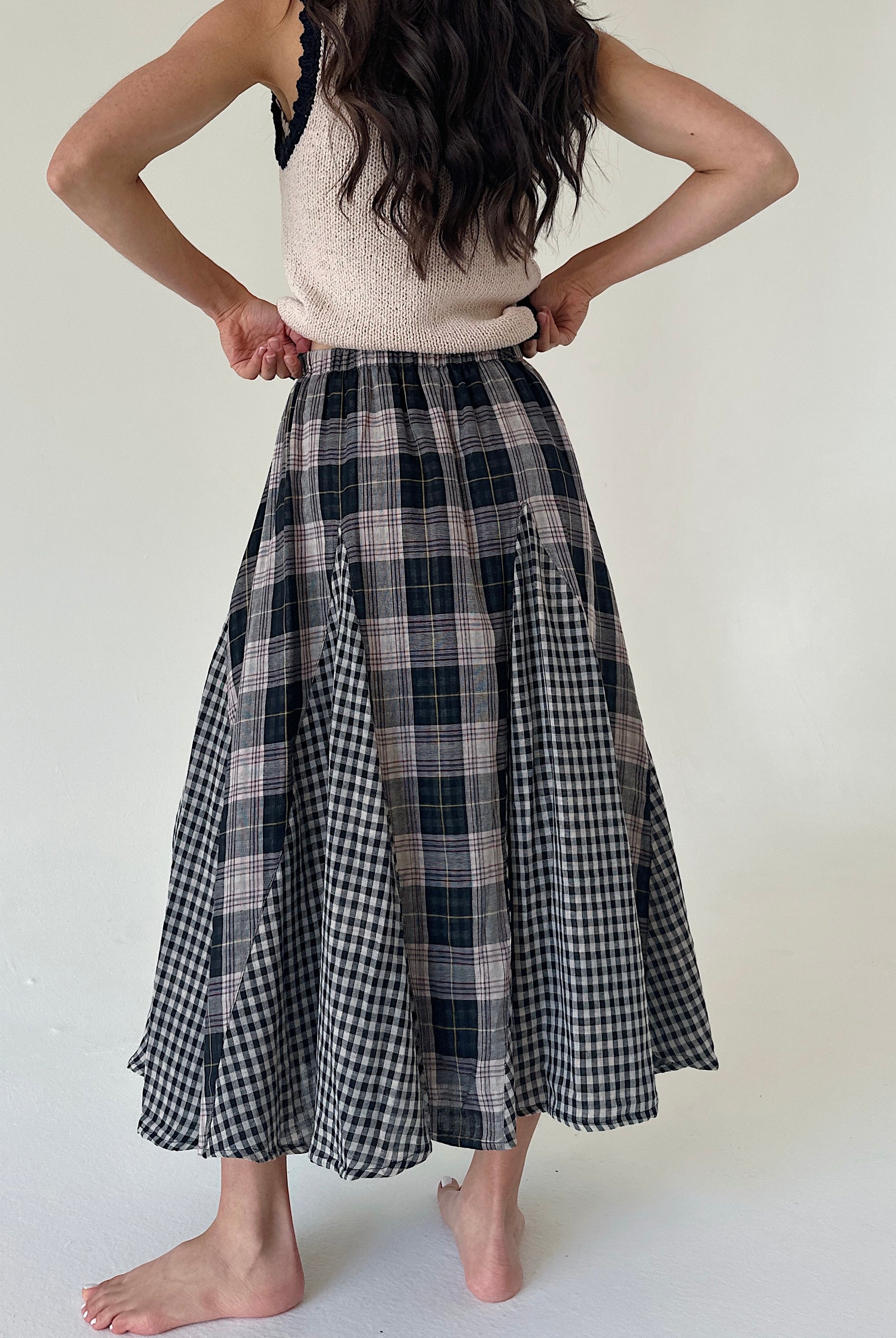 A woman shown from the back wearing a long A-line midi skirt with a mixed plaid pattern, featuring black and white gingham and dark navy/black plaid panels. She is also wearing a cream knit top, standing barefoot against a plain off-white background.