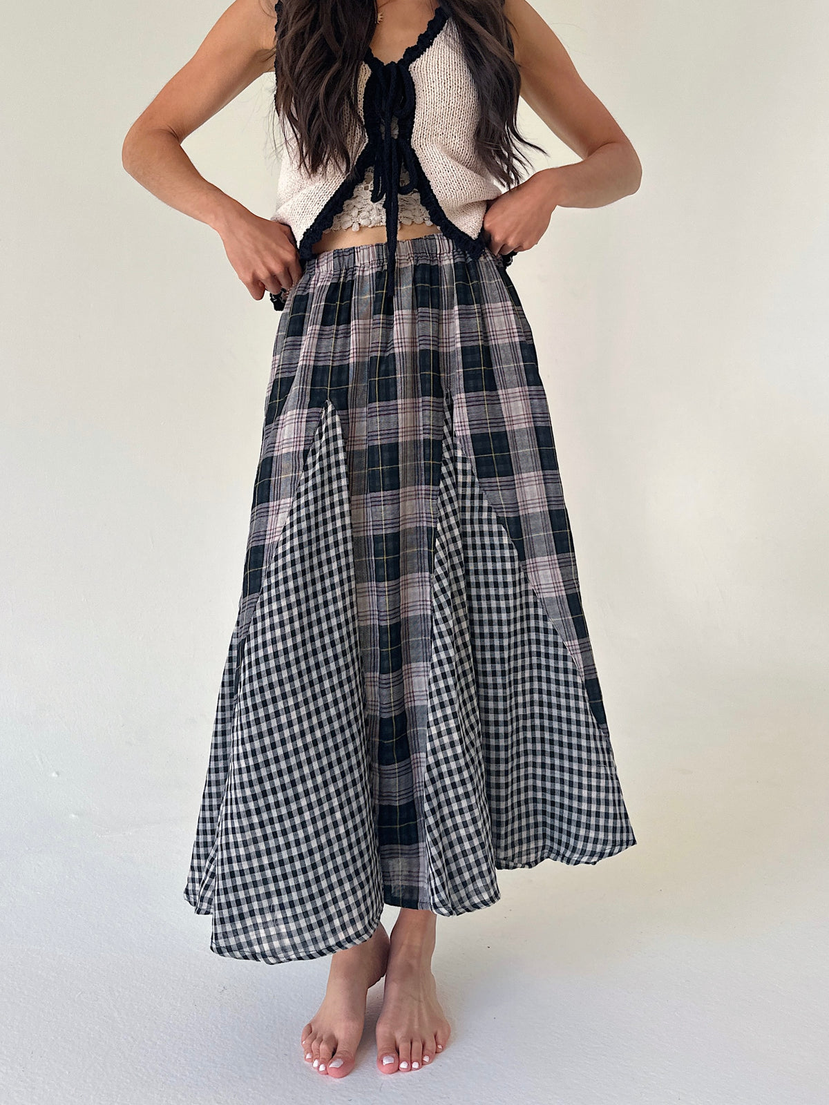 A woman wearing a long, pleated maxi skirt with a mixed plaid and gingham pattern in black, white, and muted pink, paired with a white knit halter top against a plain off-white background.