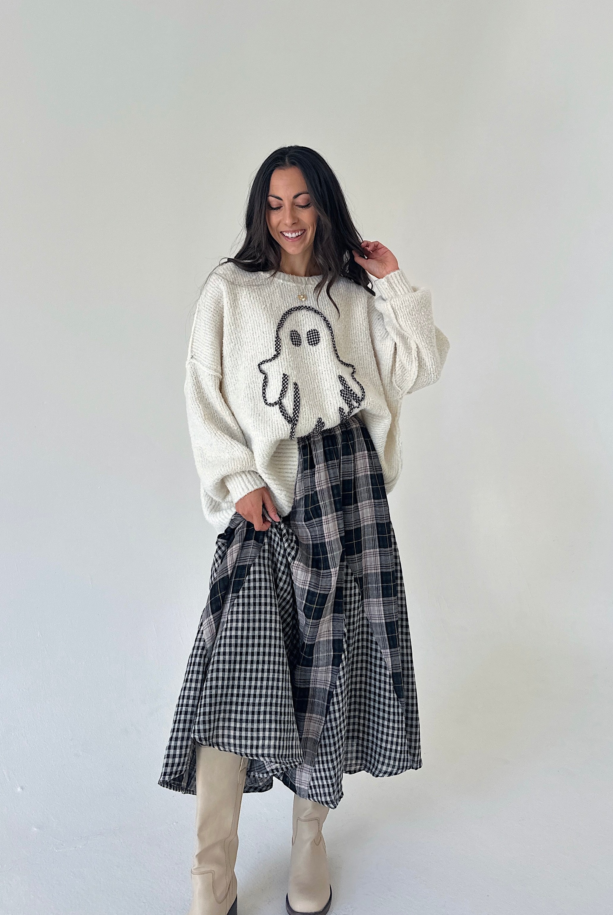 A woman stands against a plain light gray studio background wearing a cream knit sweater with a ghost graphic, a black-and-white plaid skirt, and beige knee-high boots.