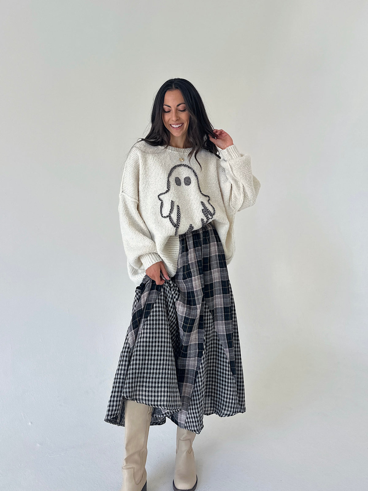 A woman stands against a plain light gray studio background wearing a cream knit sweater with a ghost graphic, a black-and-white plaid skirt, and beige knee-high boots.