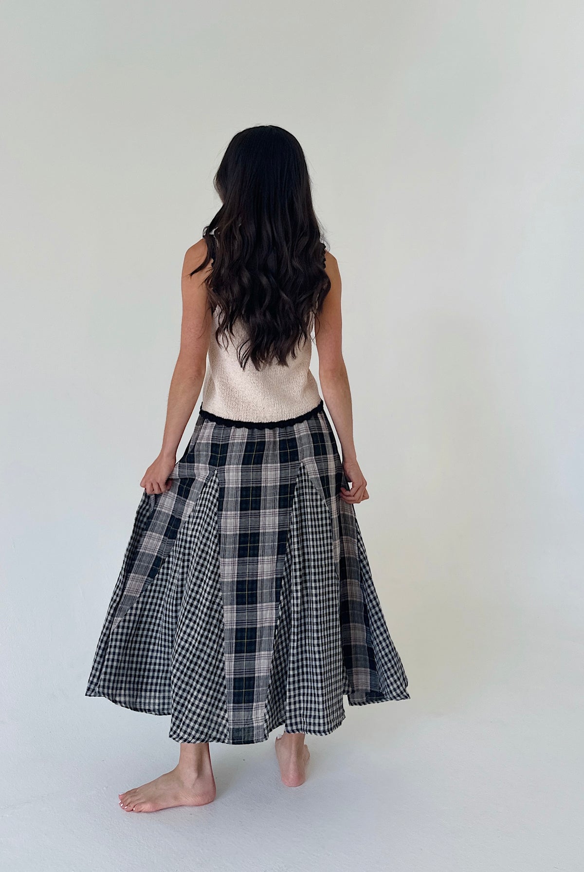 A woman viewed from behind wearing a sleeveless cream top and a black and white plaid midi skirt, standing barefoot against a plain off-white studio background.