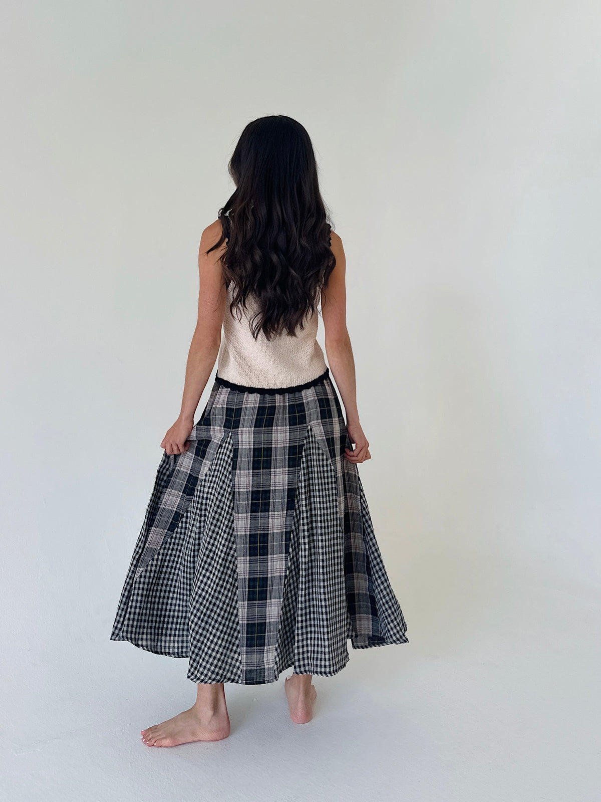A woman viewed from behind wearing a sleeveless cream top and a black and white plaid midi skirt, standing barefoot against a plain off-white studio background.