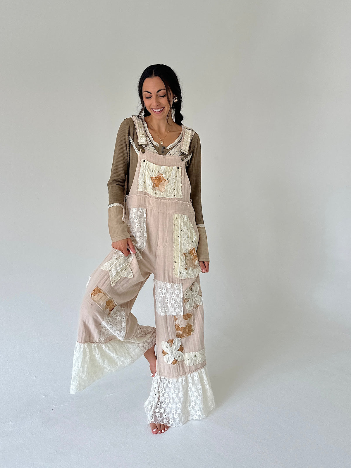 A woman modeling a bohemian-style patchwork jumpsuit in beige and brown, featuring floral lace panels and wide-leg flared pants, against a plain light gray studio background.
