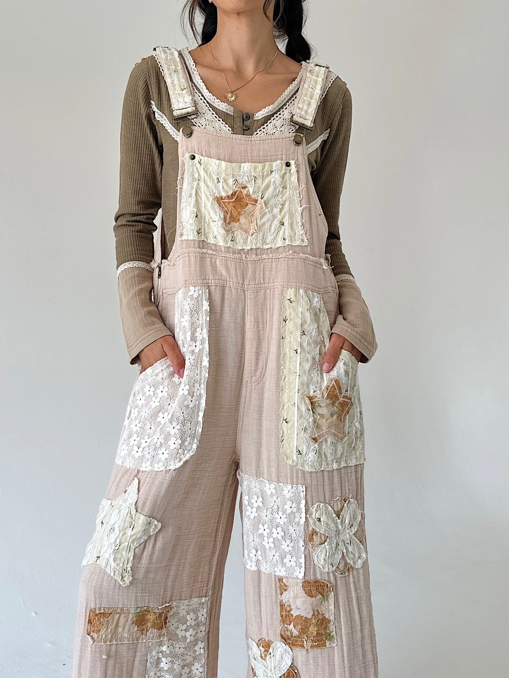 A person wearing a beige and brown patchwork overall jumpsuit with lace and floral applique details, layered over a green long-sleeve shirt, against a plain light gray background.