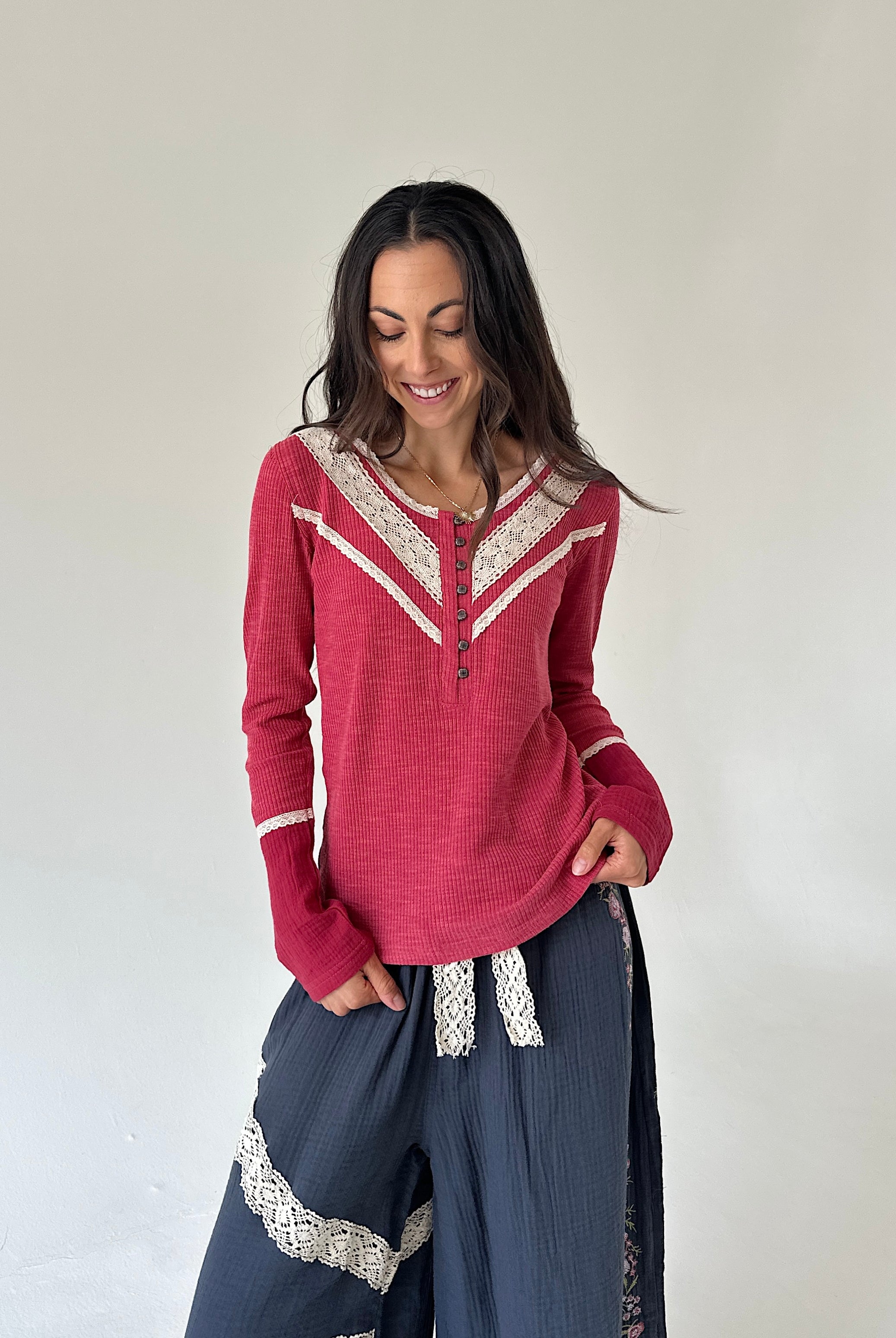 A woman modeling a long-sleeve henley top in dusty rose pink with cream lace trim, paired with dark blue wide-leg pants with lace details, against a plain off-white wall.