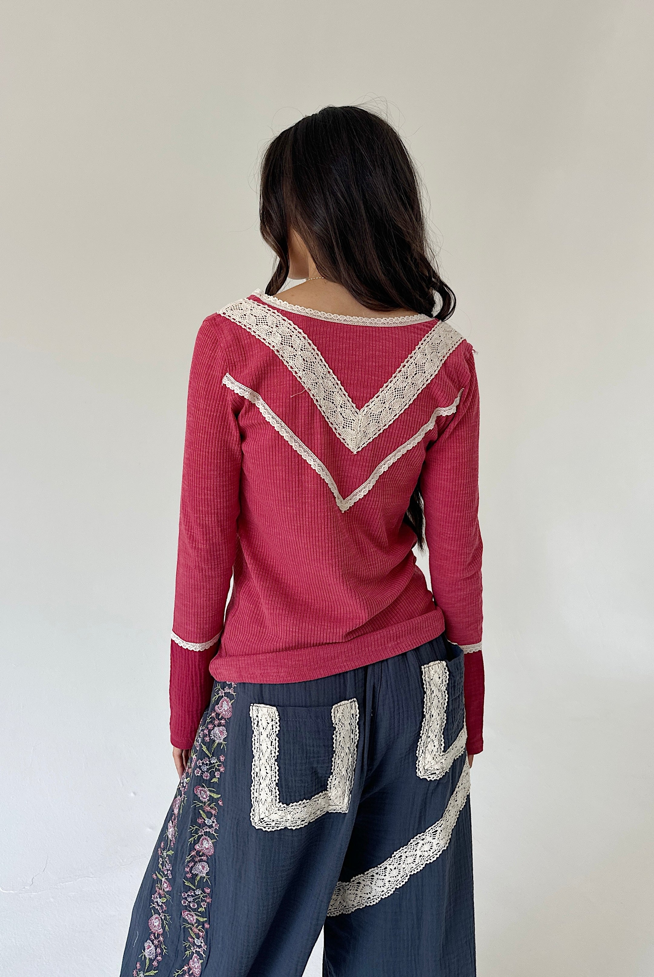 A woman shown from behind wearing a dusty rose long-sleeve knit top with cream lace trim and dark blue wide-leg pants with floral and lace details, set against a plain off-white wall.