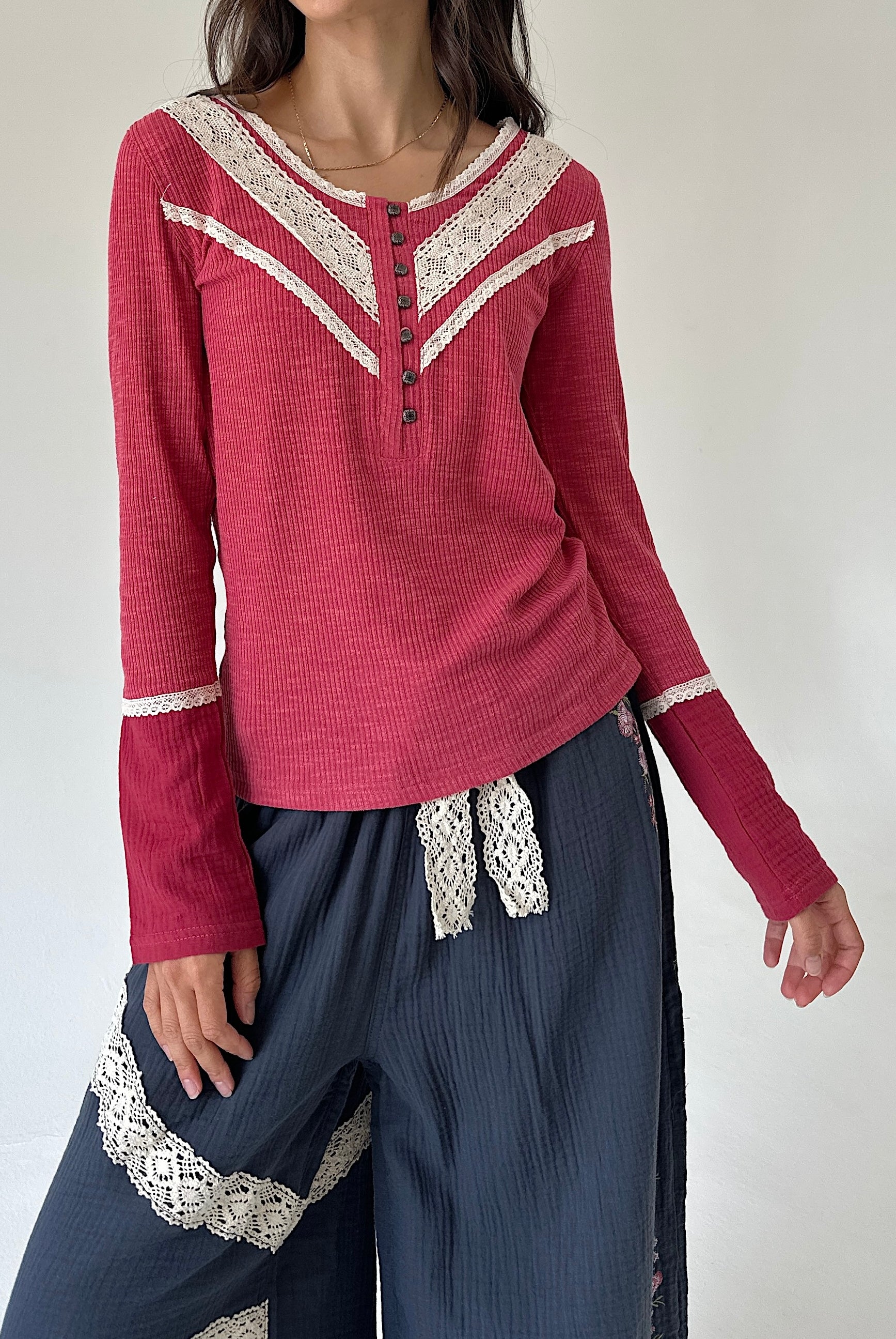 A woman wearing a long-sleeve knit top in dusty rose pink with cream lace trim along the V-neckline and cuffs, paired with dark blue wide-leg pants featuring cream lace details.