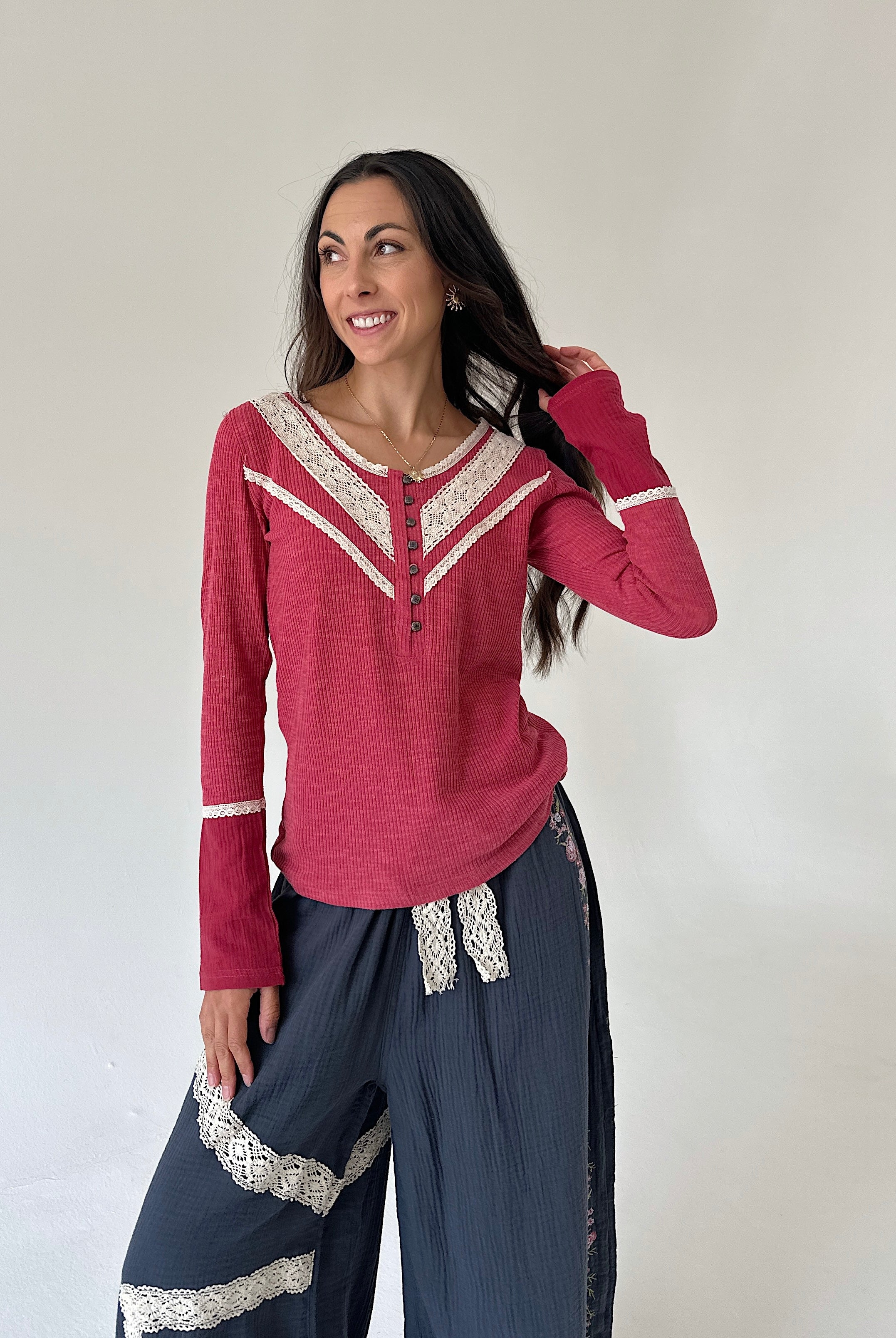 A woman modeling a long-sleeve knit top in dusty rose pink with cream lace trim, paired with dark blue gauze pants featuring lace accents, against a plain off-white wall.