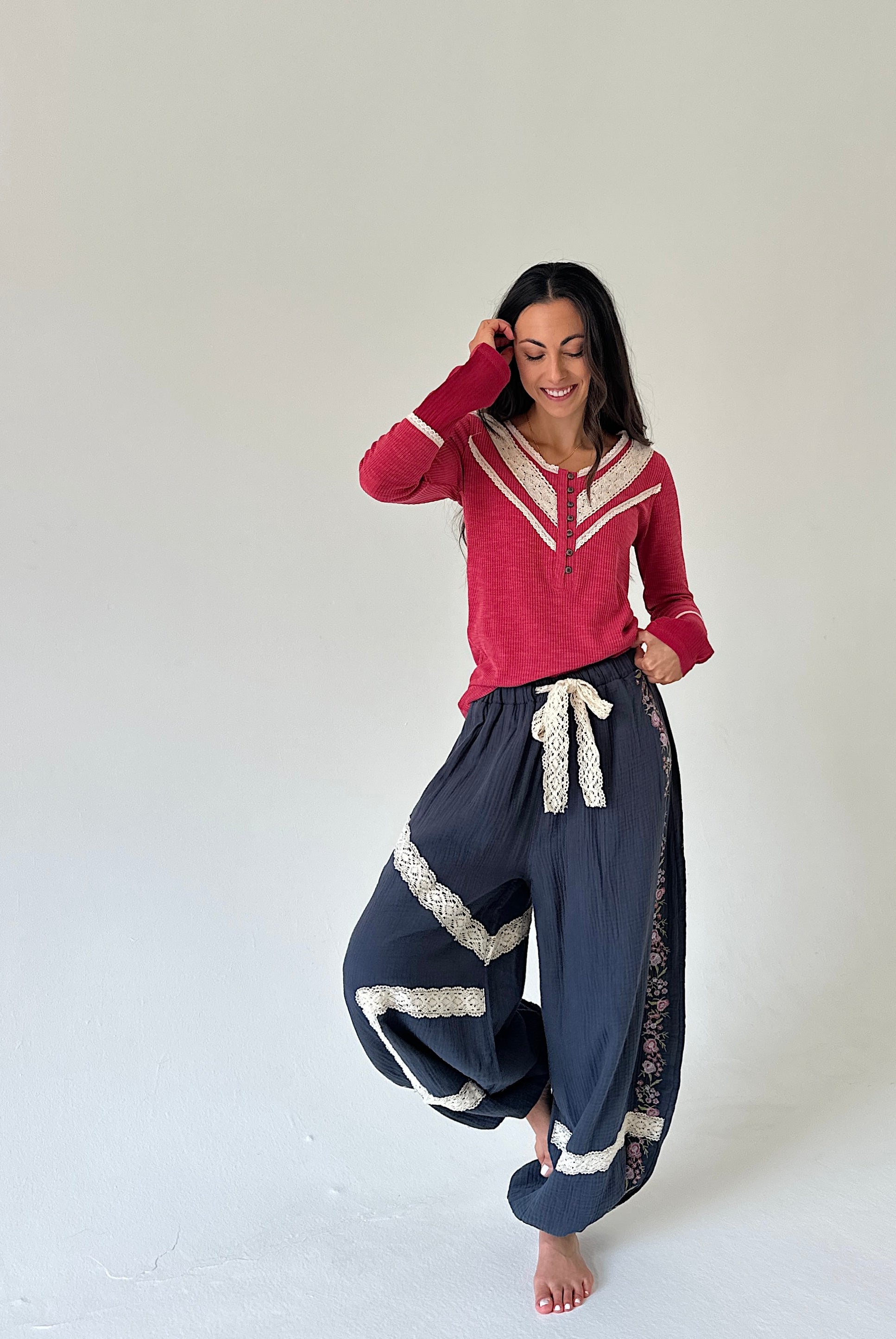 A woman modeling a two-piece outfit consisting of a red long-sleeve henley top with cream trim and navy blue harem-style pants with cream lace trim and a cream drawstring, set against a plain off-white studio background.