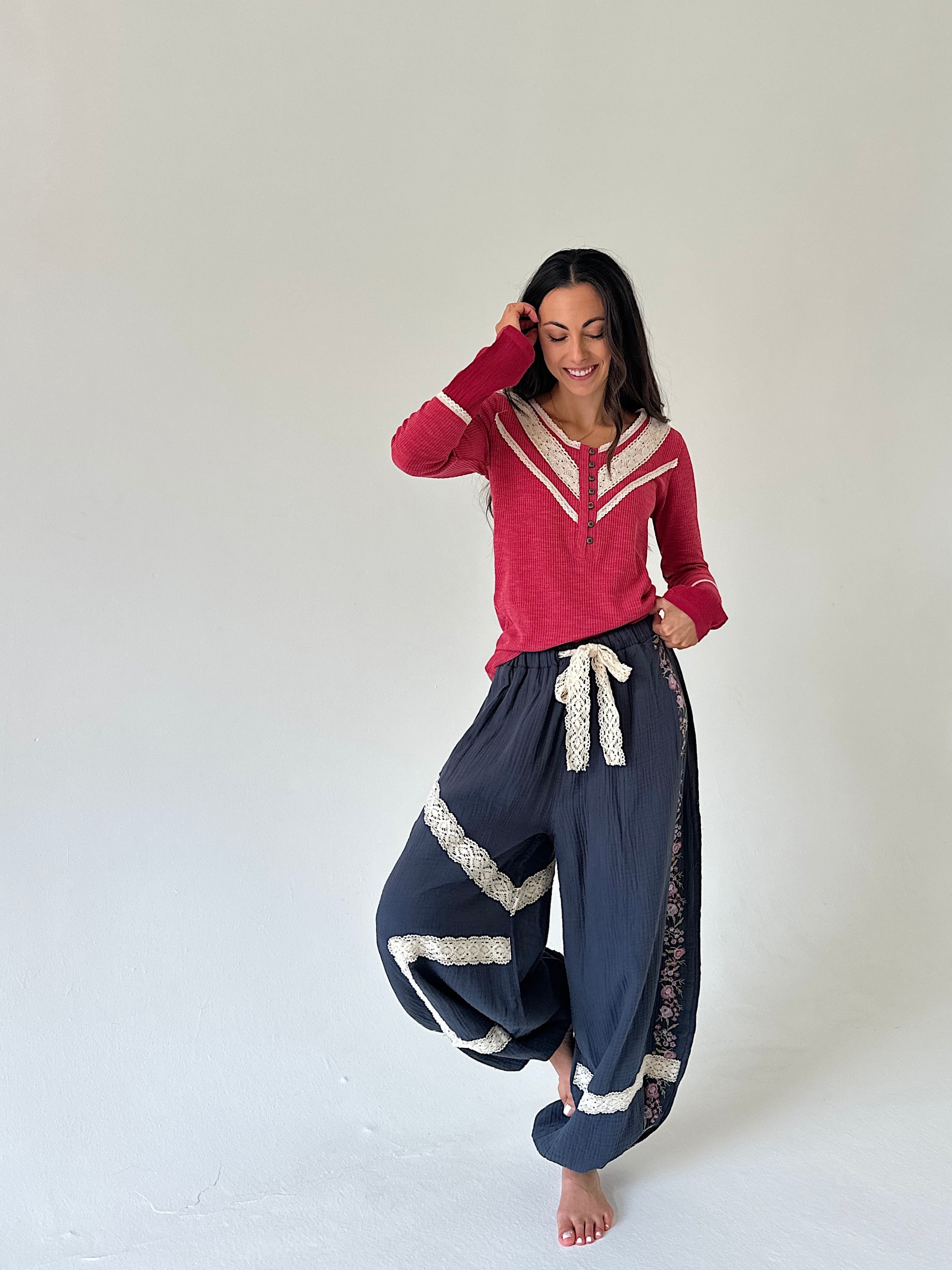 A woman modeling a two-piece outfit consisting of a red long-sleeve henley top with cream trim and navy blue harem-style pants with cream lace trim and a cream drawstring, set against a plain off-white studio background.