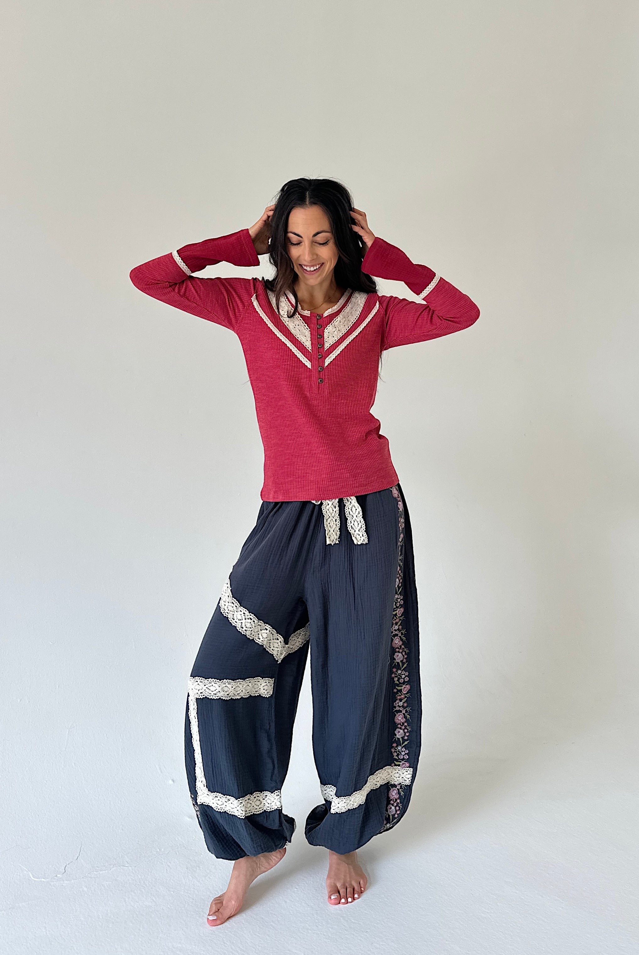 A woman modeling a red long-sleeve henley shirt with white trim and navy blue harem pants with white lace details, standing against a plain light gray studio background.