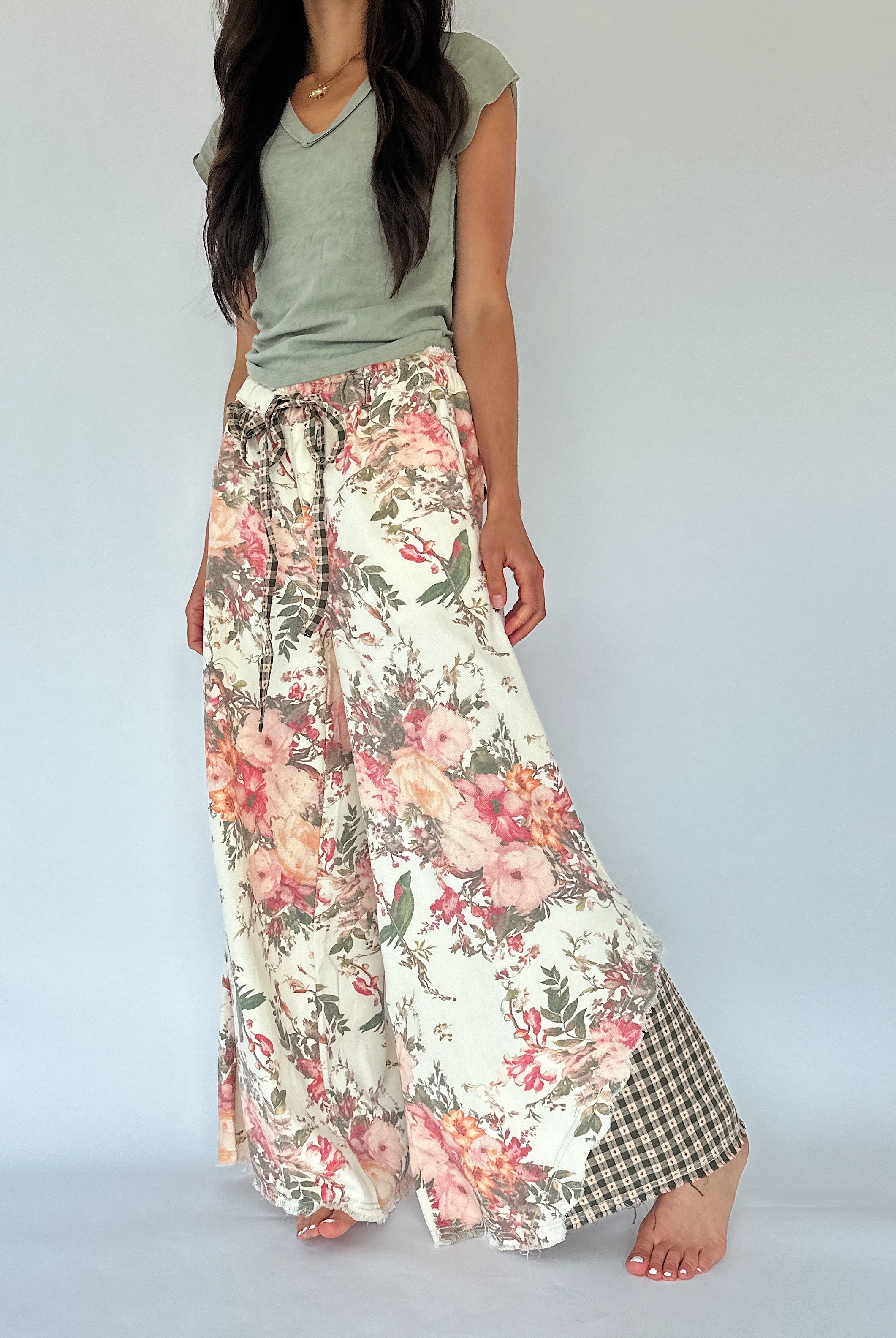 A woman modeling wide-leg floral pants with a cream base, pink and red flowers, green leaves, and a green-and-white gingham check panel at the hem, paired with an olive green top against a light gray studio background.