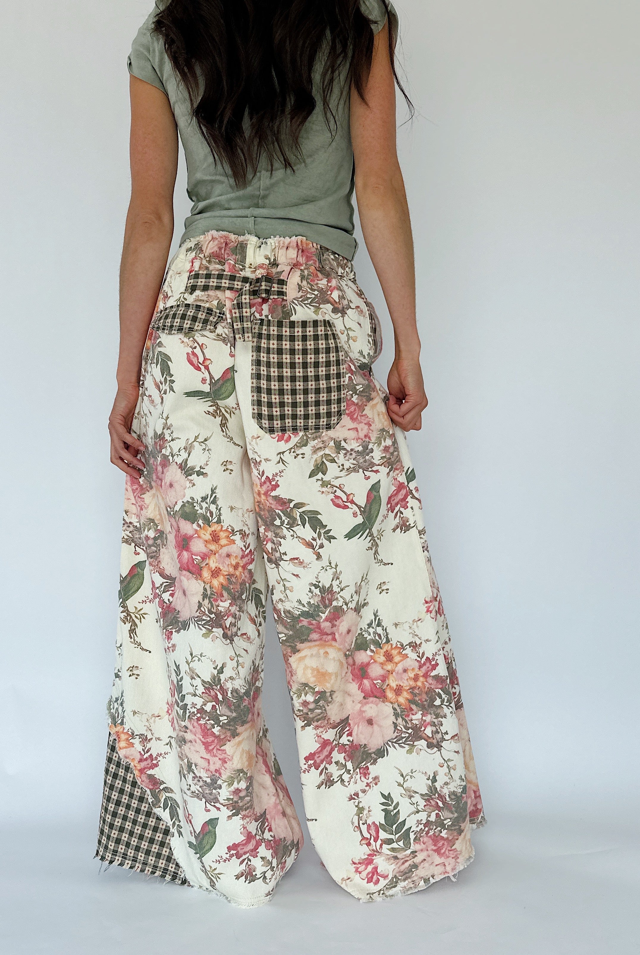 A person shown from the back wearing wide-leg floral pants with gingham patch pockets and a sage green top, photographed against a plain light gray studio background.