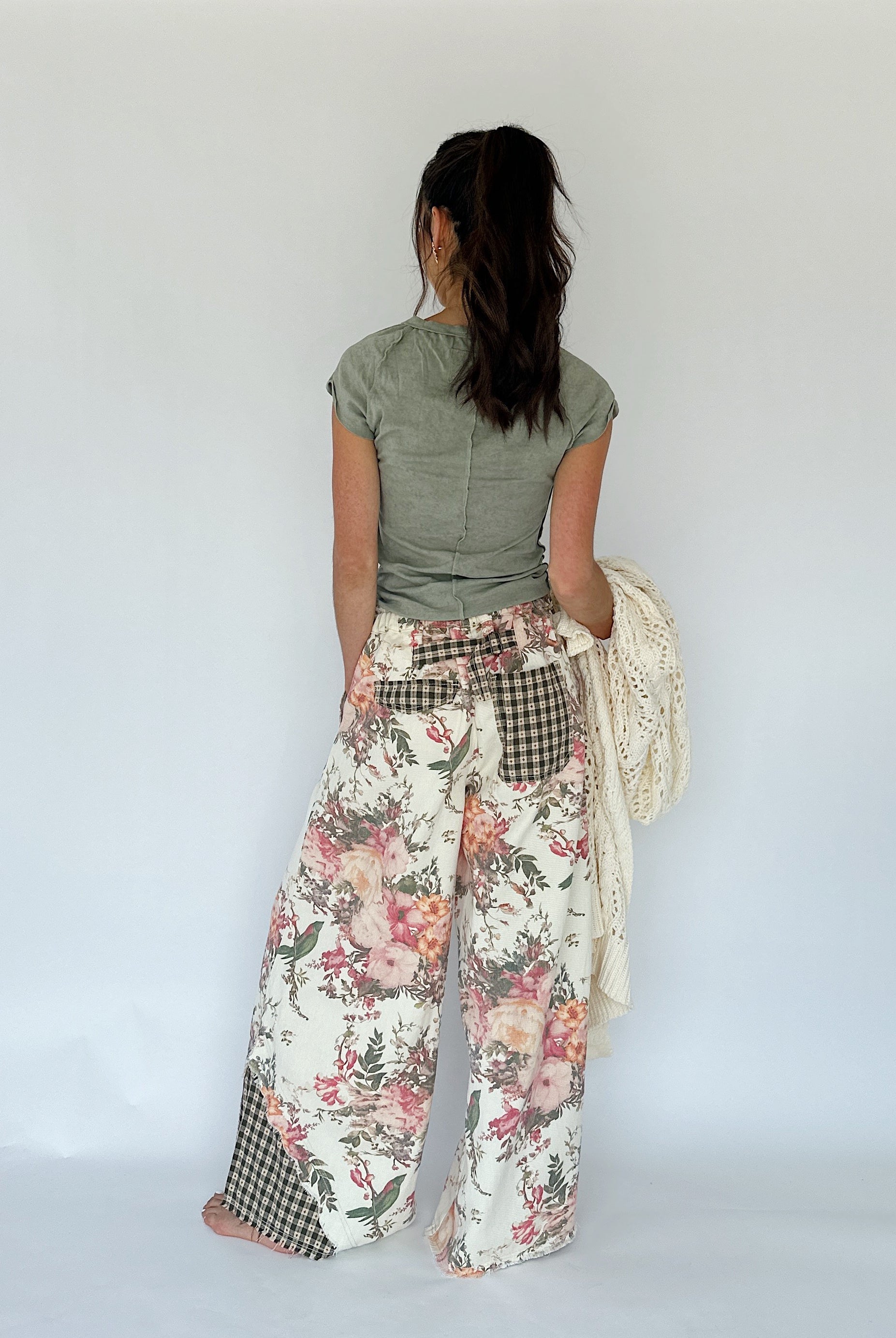A woman shown from behind wearing wide-leg patchwork pants with a floral print, gingham pockets, and a green top, holding a cream knit sweater against a plain white studio background.