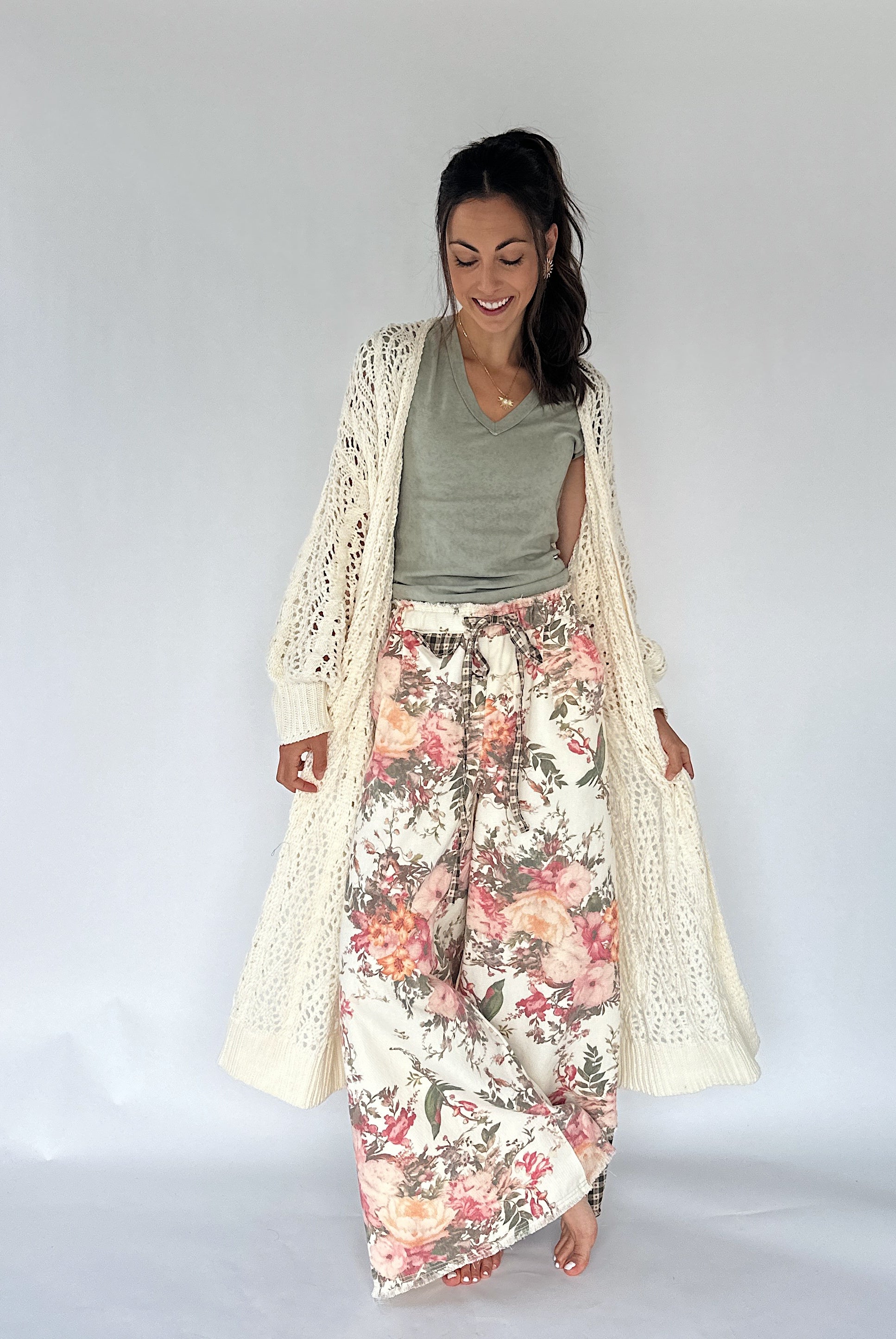 A woman modeling a bohemian-style outfit consisting of a cream open-front crochet cardigan, a sage green V-neck top, and wide-leg floral print pants with pink, red, and green flowers on a cream background.