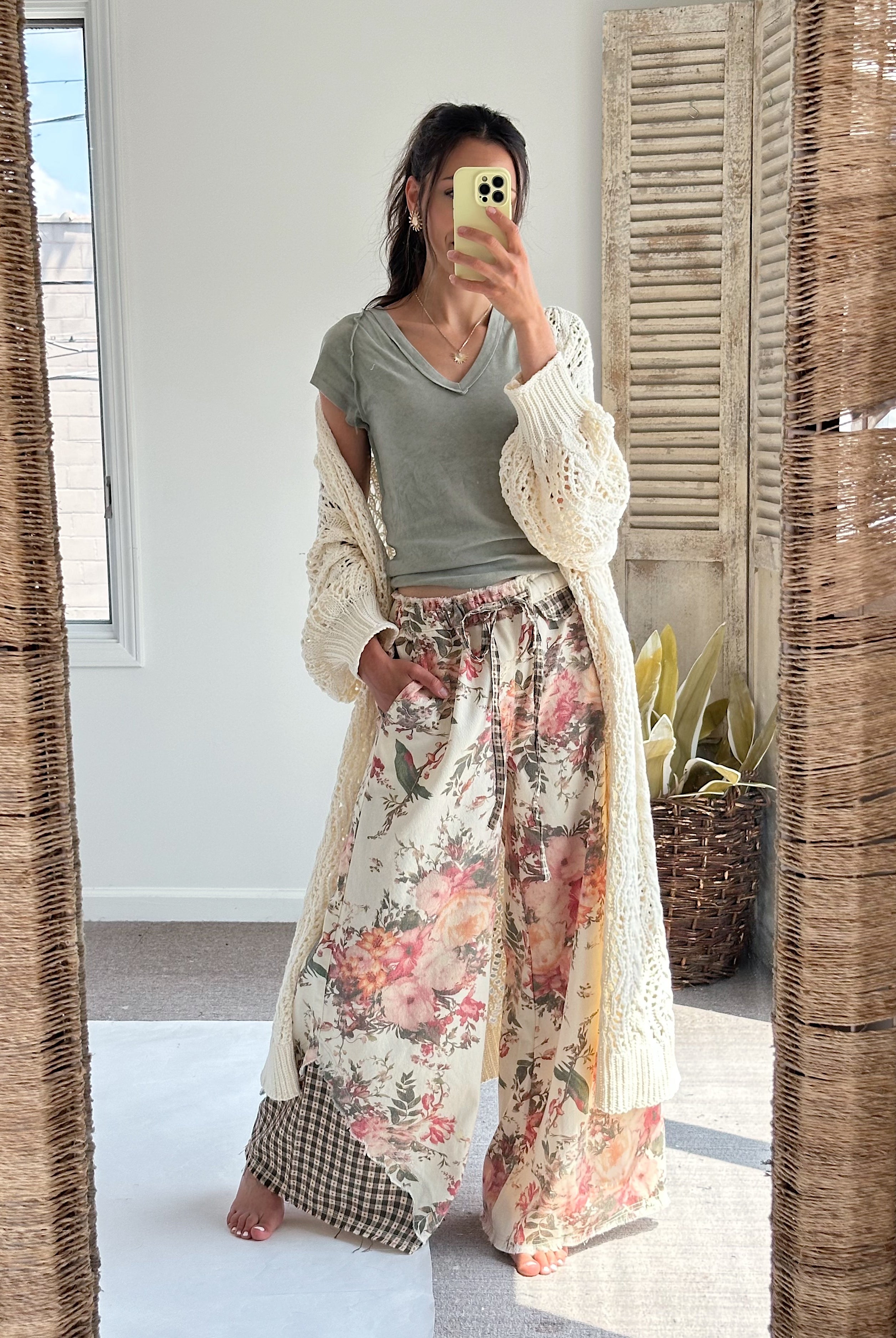 A woman takes a mirror selfie wearing a bohemian-style outfit consisting of a sage green V-neck t-shirt, a cream open-knit cardigan, and wide-leg floral print pants with a gingham leg overlay.
