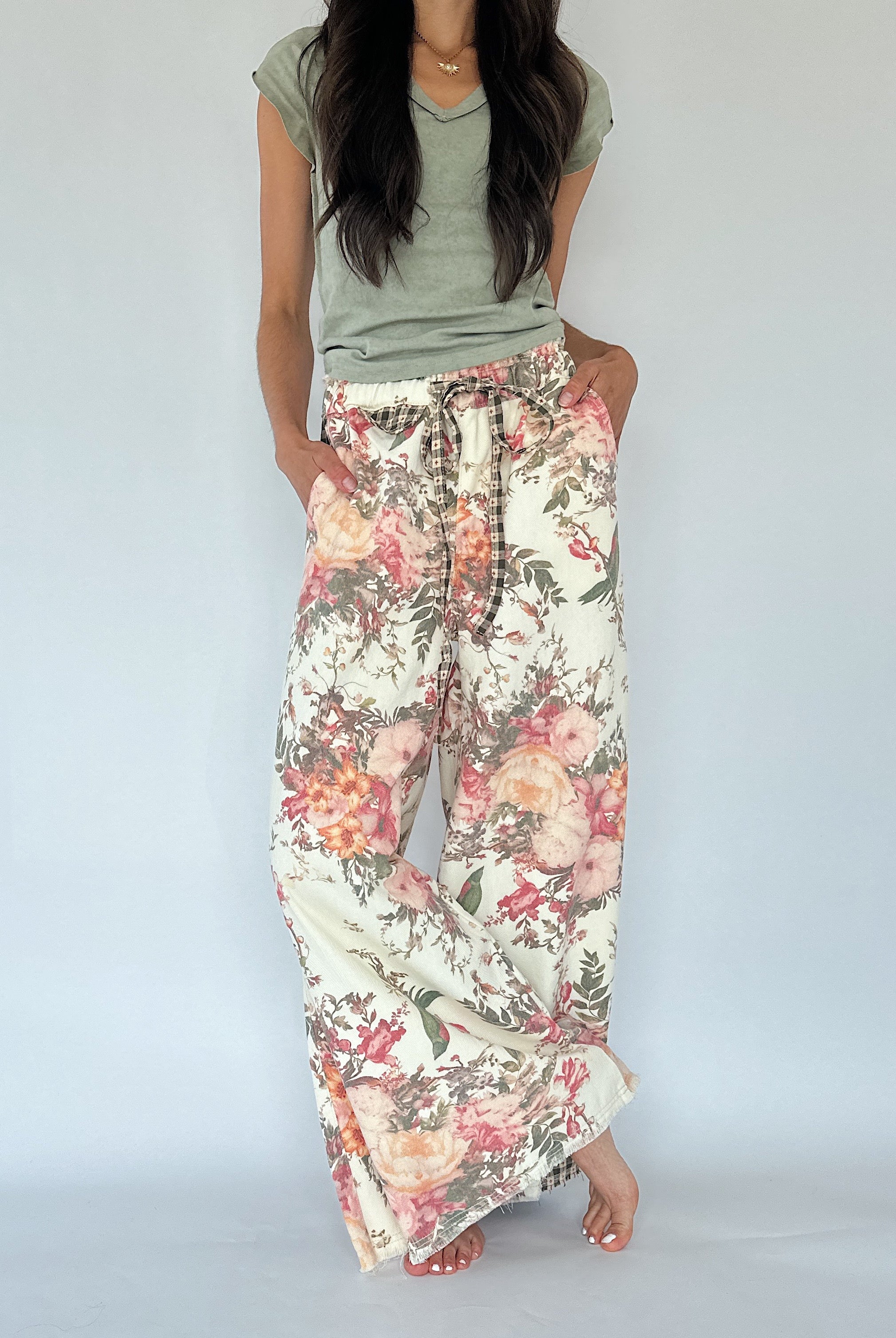 A woman wearing a sage green t-shirt and wide-leg floral pants with a pink, orange, and green botanical print on a cream background, standing against a light gray studio backdrop.