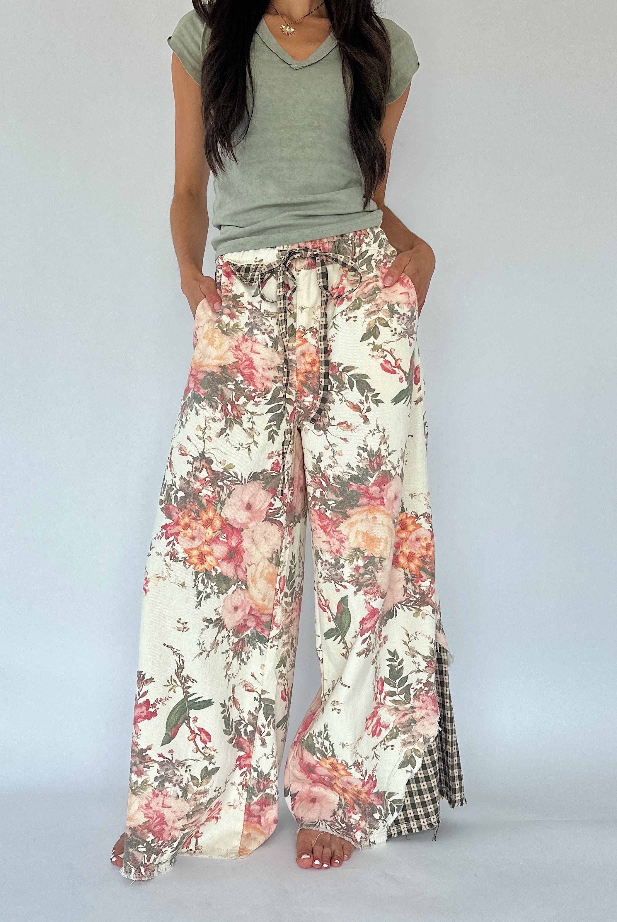 A model wearing wide-leg floral print pants with a drawstring waist and a green-and-white gingham side panel, paired with a sage green V-neck t-shirt against a plain light gray background.