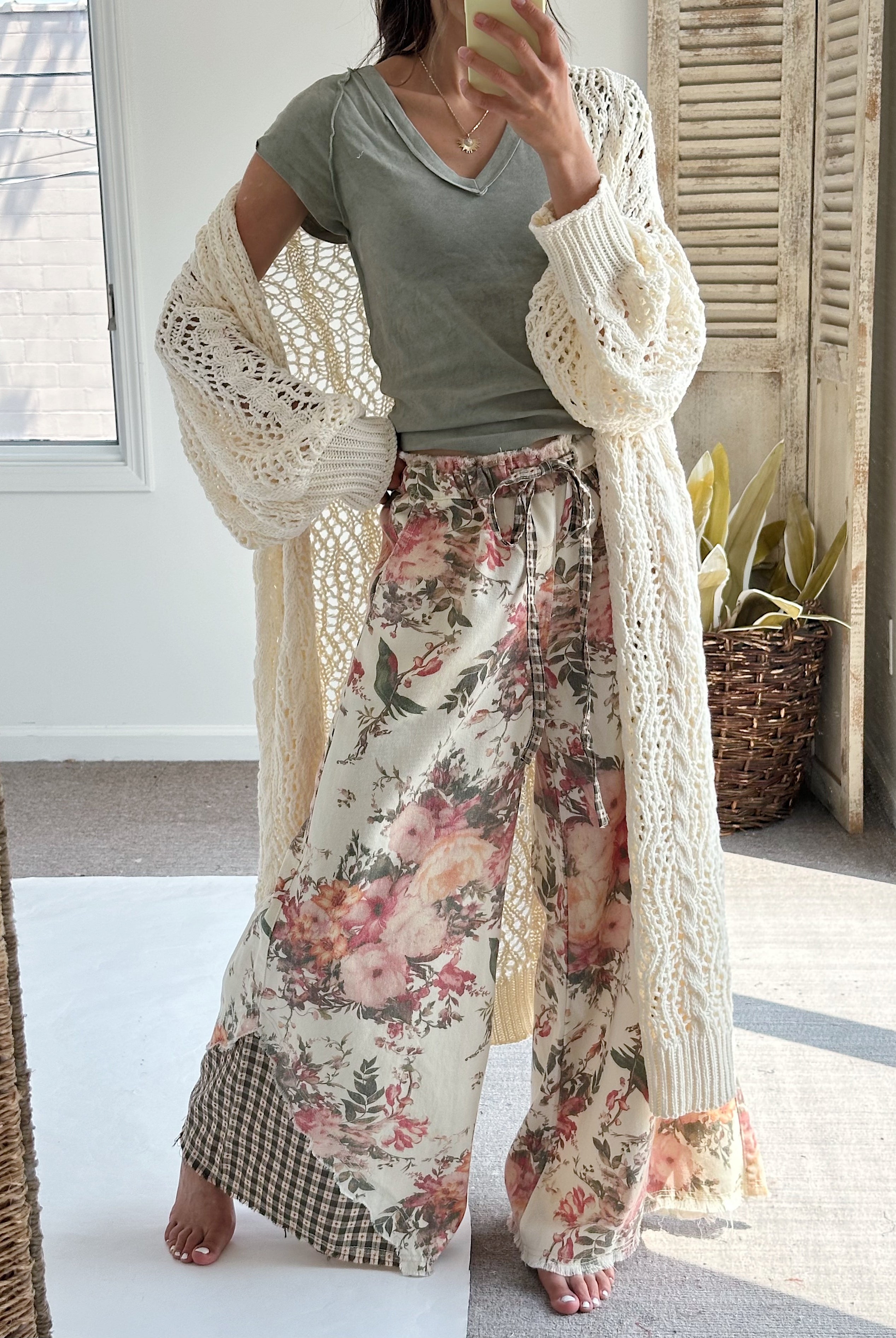 A woman takes a mirror selfie wearing a sage green t-shirt, a cream open-knit cardigan, and wide-leg floral pants with a gingham hem patch. The setting is a bright room with a window, shutters, and a potted plant.
