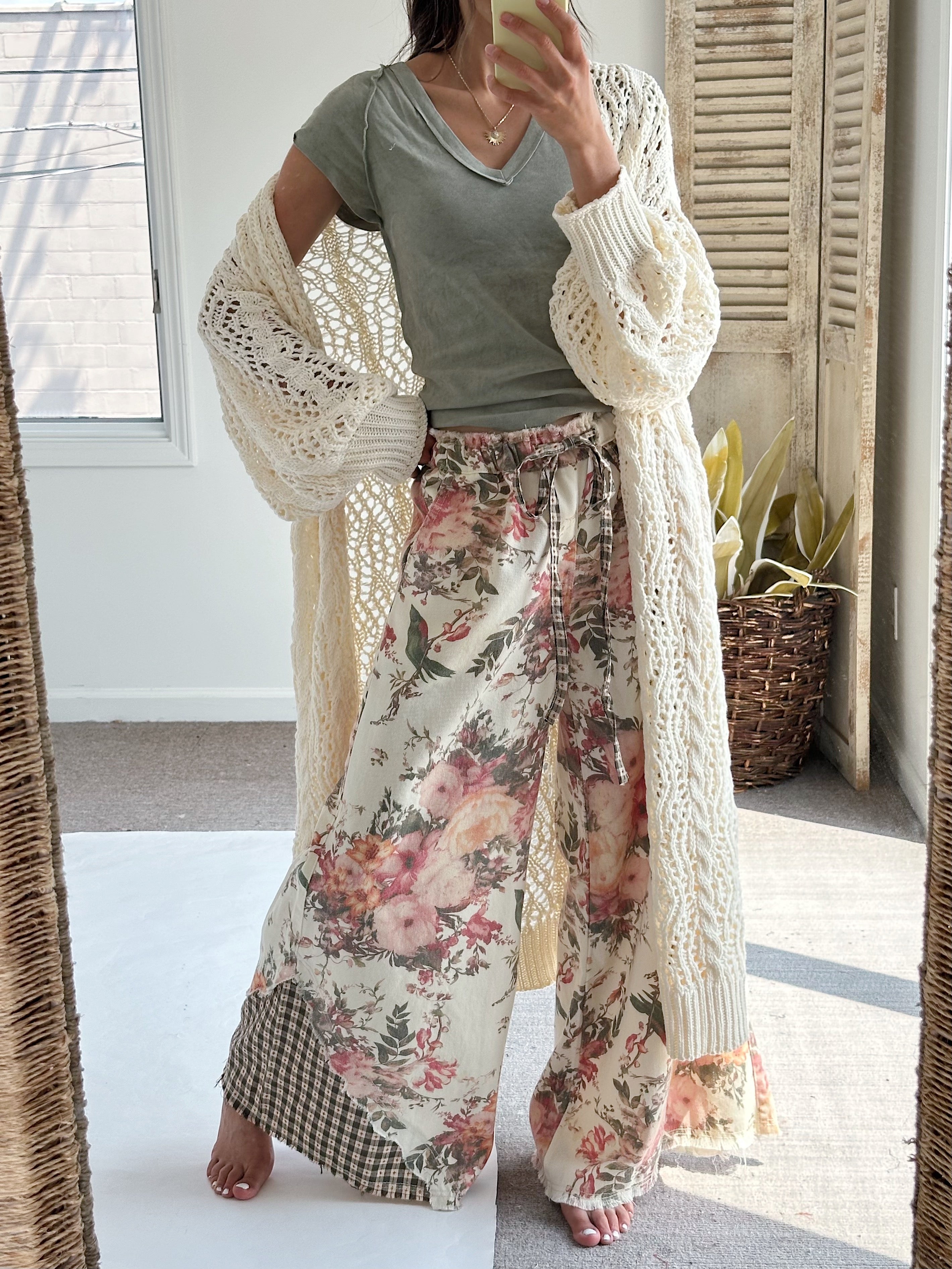 A woman takes a mirror selfie wearing a sage green t-shirt, a cream open-knit cardigan, and wide-leg floral pants with a gingham hem patch. The setting is a bright room with a window, shutters, and a potted plant.
