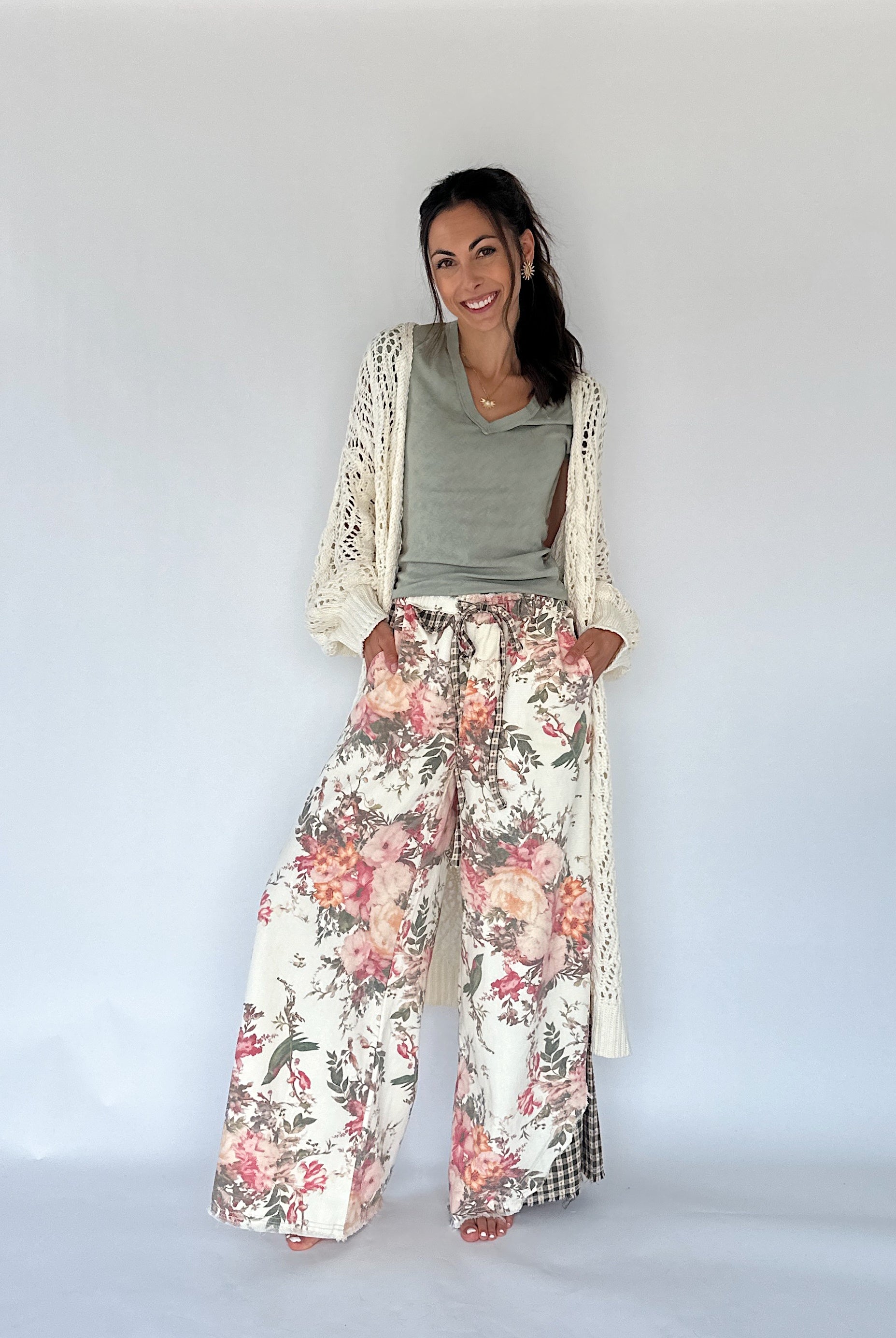 A woman modeling a bohemian-style outfit consisting of wide-leg floral print pants, a sage green top, and a cream open-knit cardigan, set against a plain white studio background.
