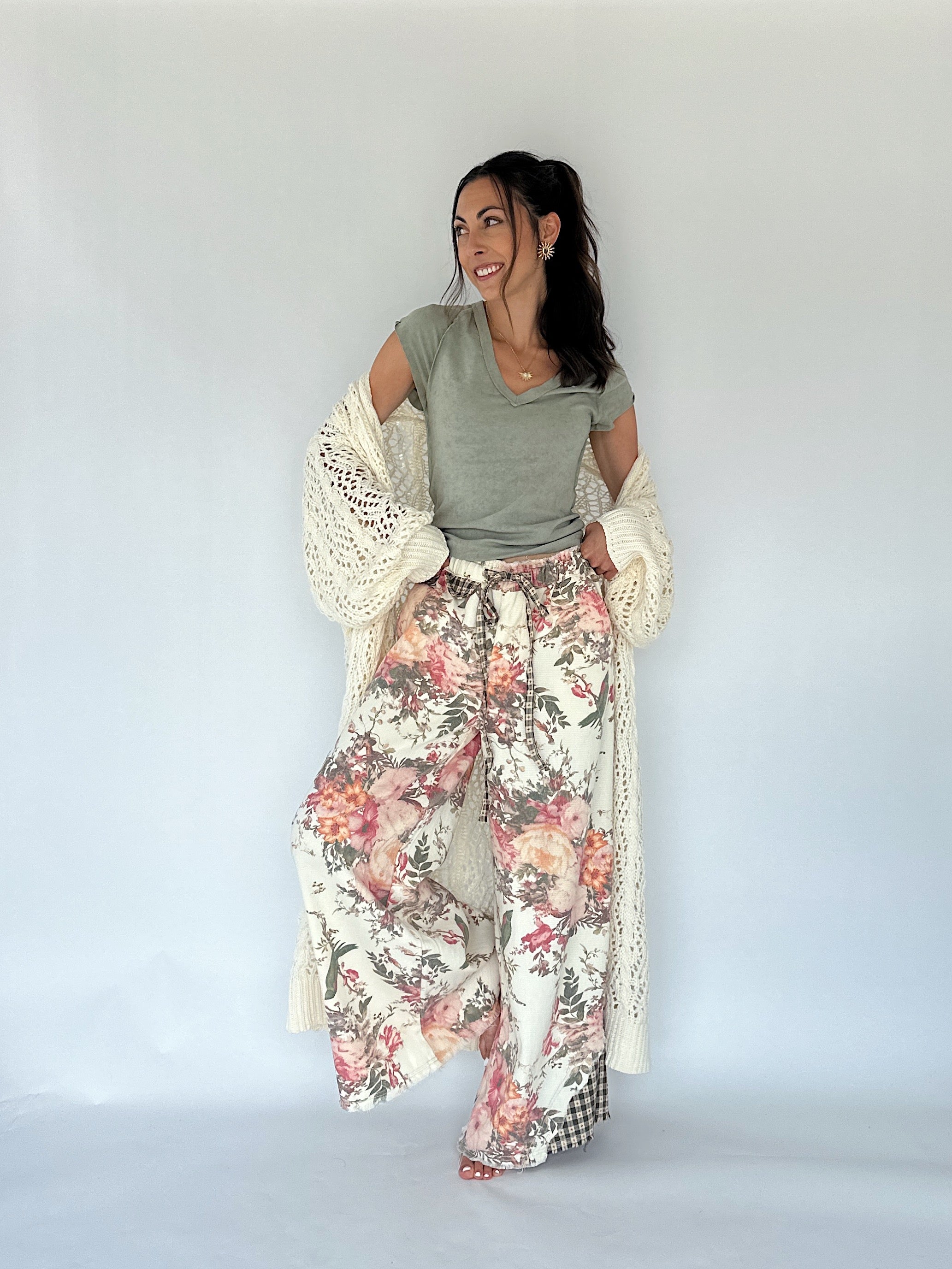 A woman modeling a bohemian-style outfit consisting of a sage green top, wide-leg floral print pants, and a cream crochet cardigan, set against a plain light gray studio background.