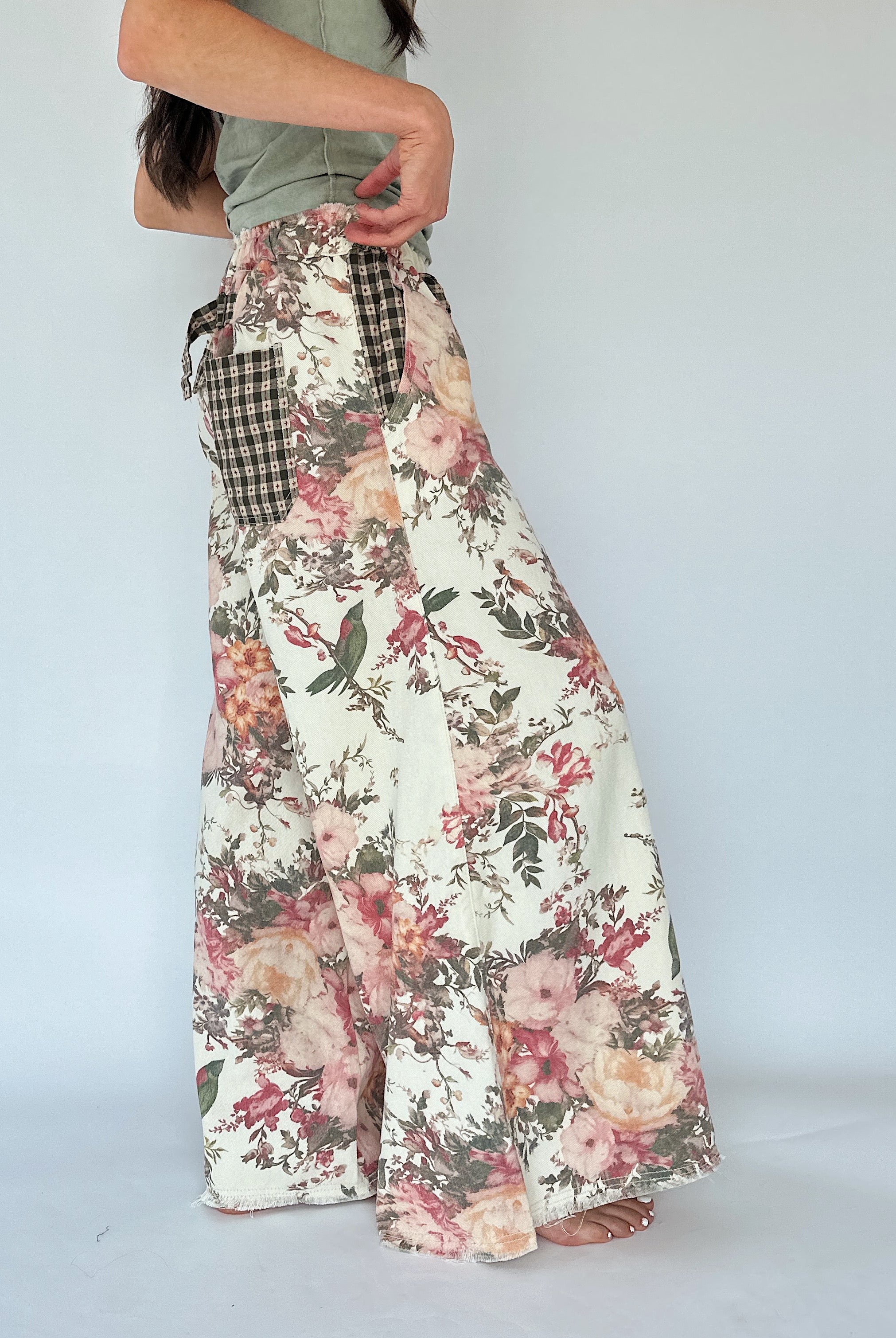 A person from the side wearing a pair of wide-leg floral pants with a gingham tie detail at the waist, shown from the torso down against a plain light gray studio background.