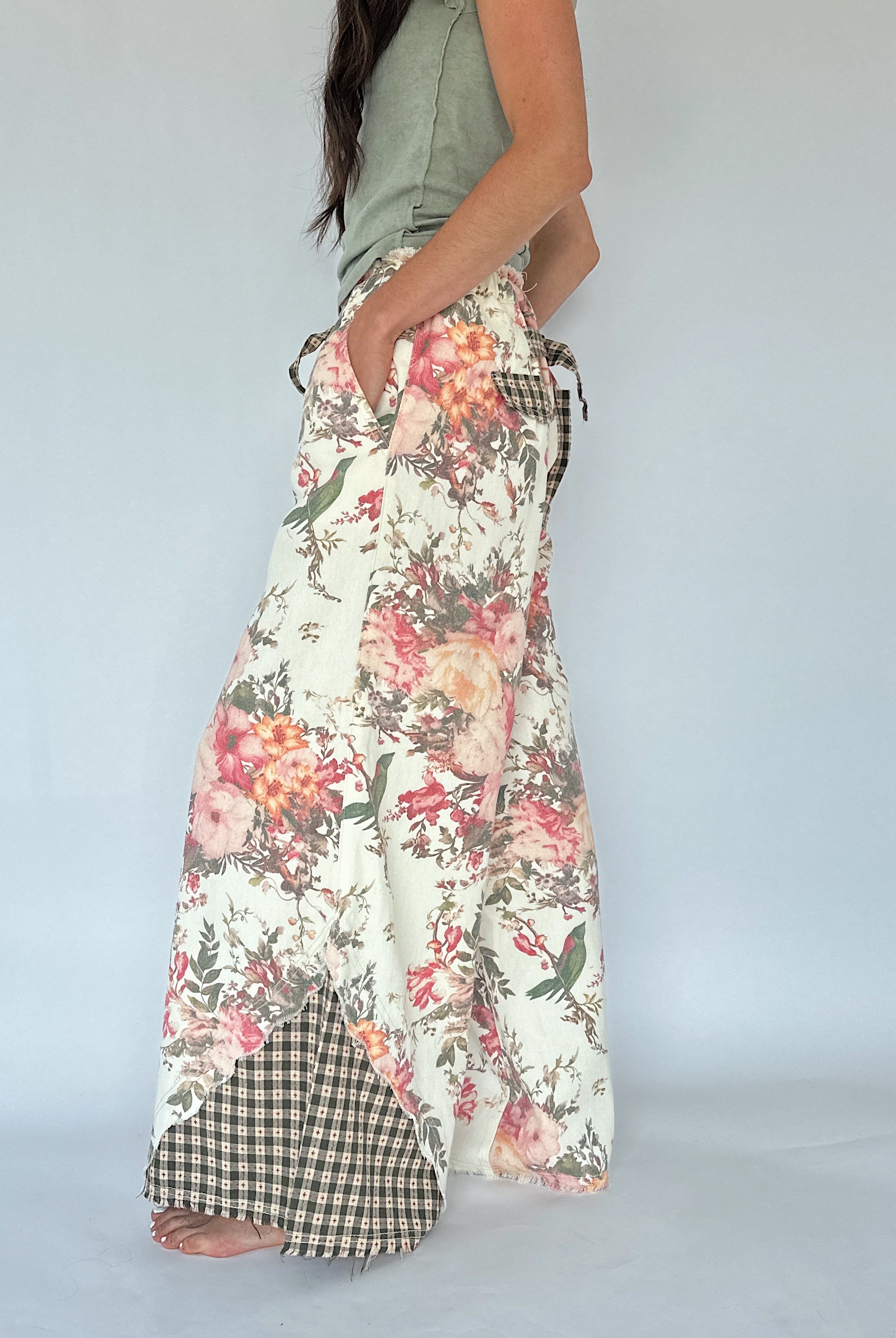A woman wearing long, wide-leg pants with a floral print on a cream background, featuring a green and white gingham checkered hem and side pocket detail, paired with a sage green sleeveless top against a light gray studio background.