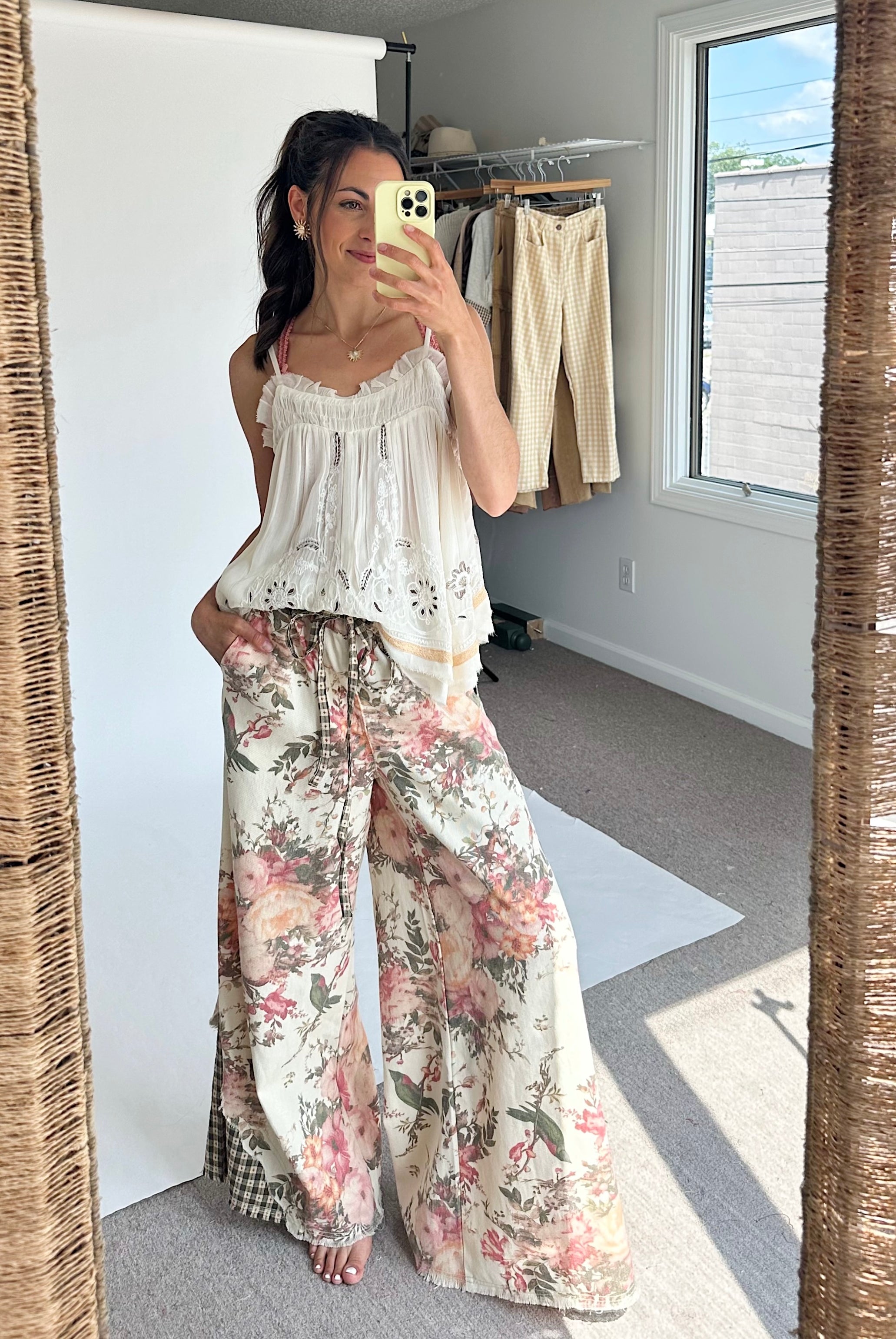 A woman takes a mirror selfie in a room, wearing a white sleeveless lace-trim camisole top and wide-leg floral pants with pink, green, and beige tones.