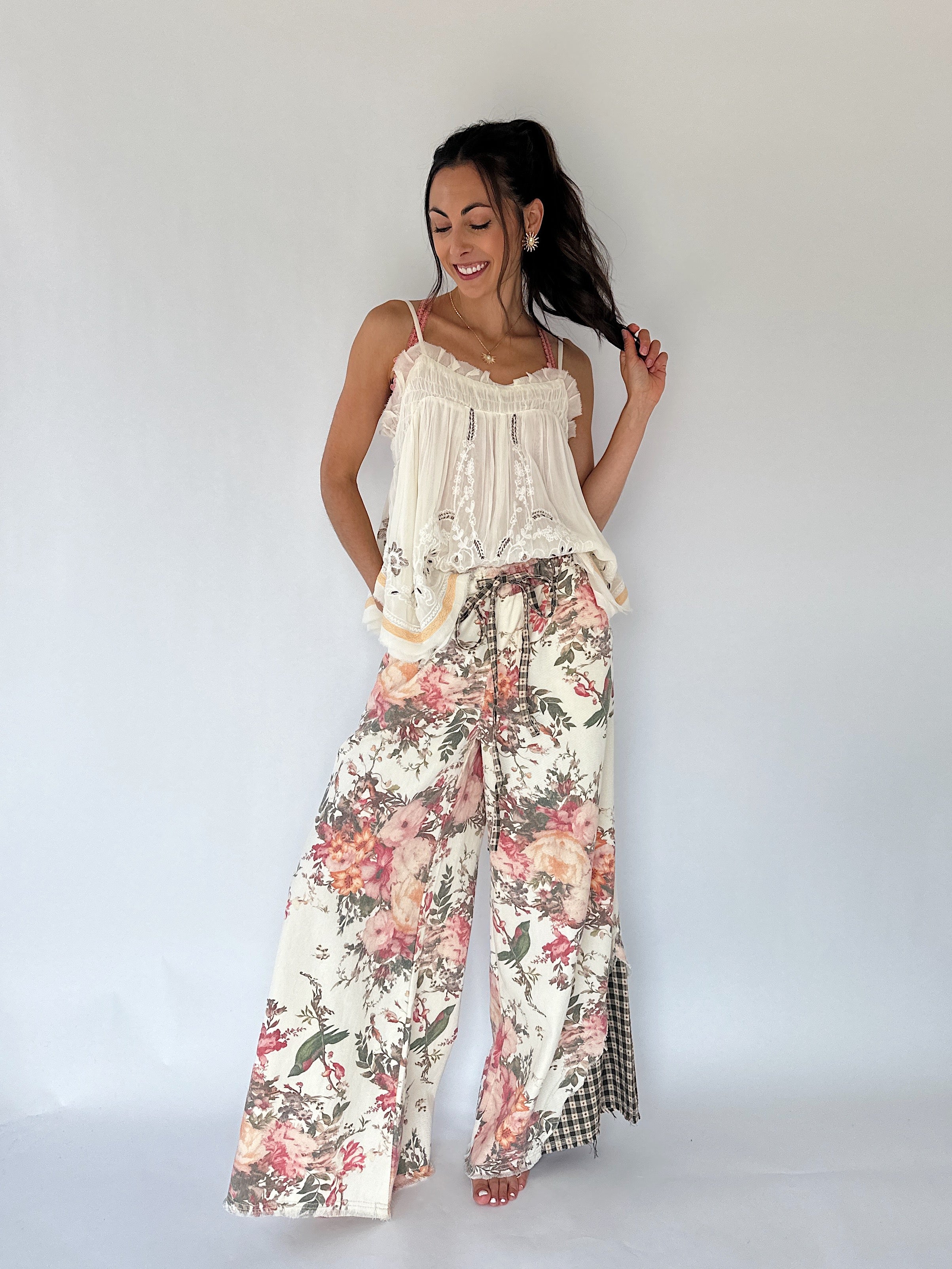 A woman modeling a bohemian-style two-piece outfit featuring a cream lace-trimmed camisole top and matching wide-leg pants with a pink, red, and green floral print, set against a plain white studio background.