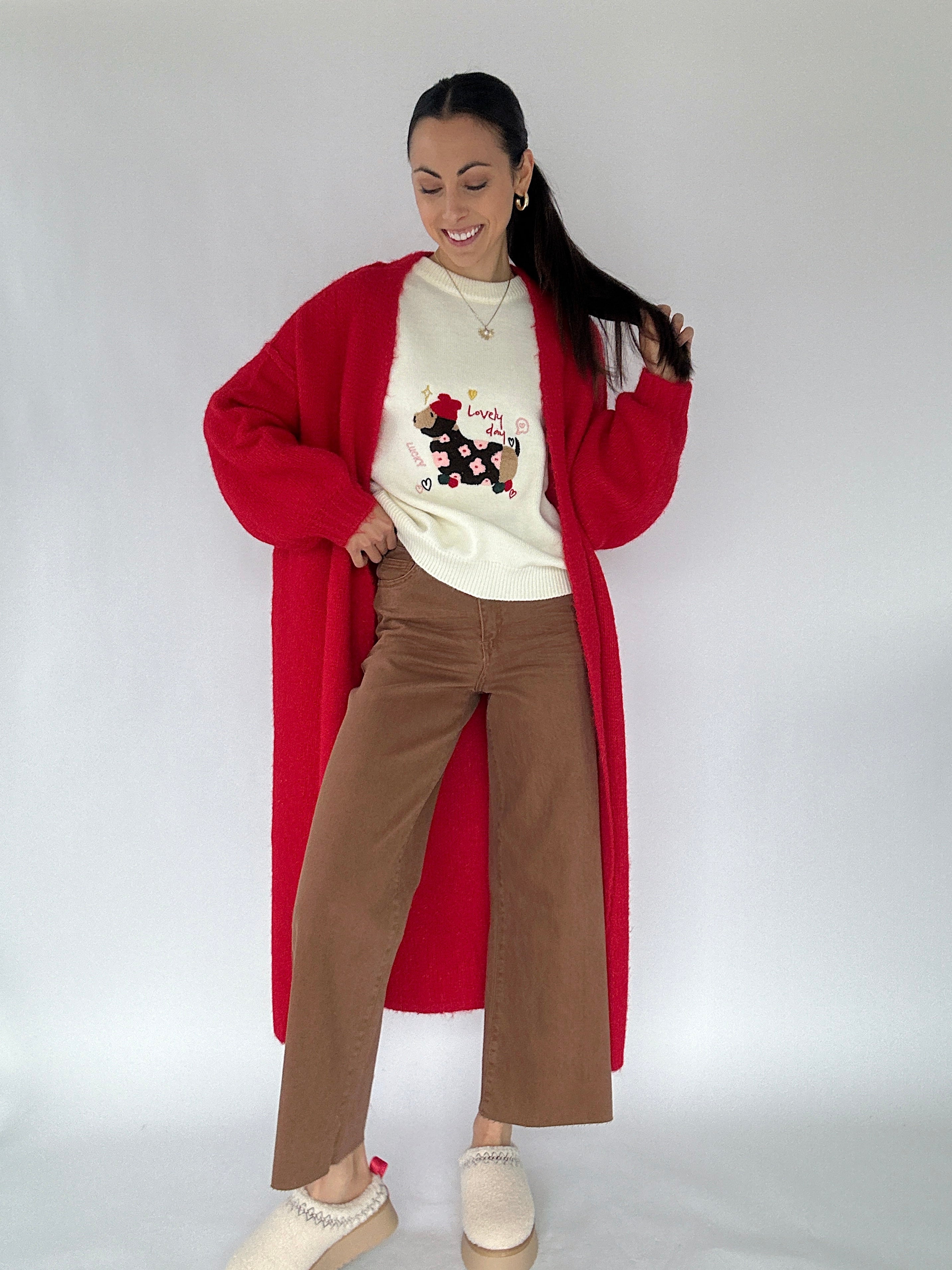 Woman wearing a long red cardigan, white sweater with graphic dog design, and brown pants on a plain background