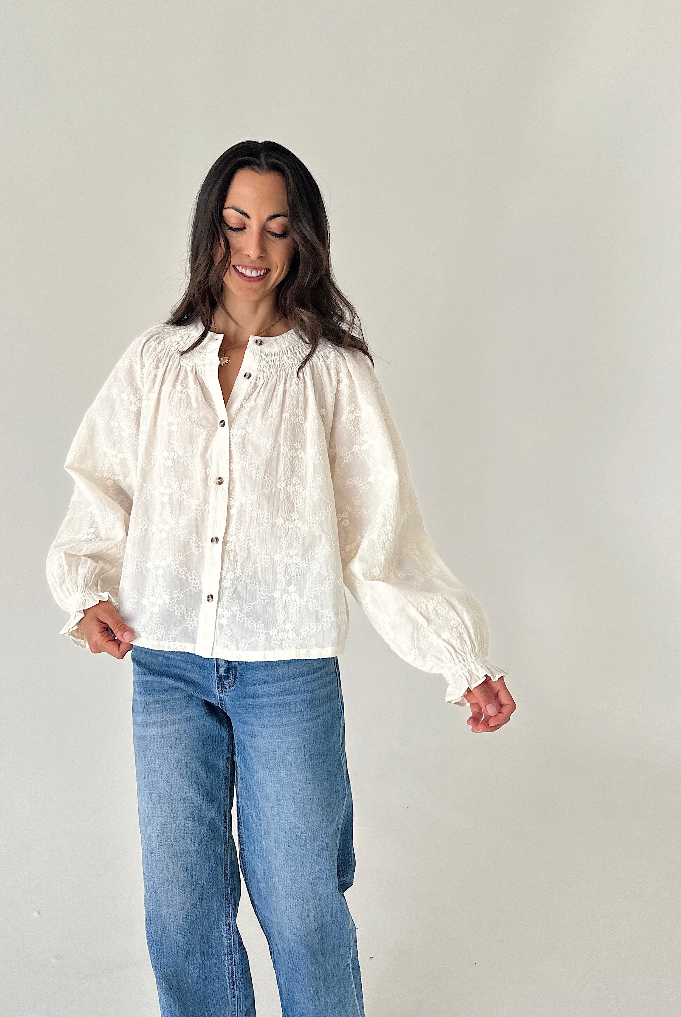A woman wearing a cream-colored, textured long-sleeve blouse with gathered cuffs and a button-down front, paired with blue jeans, standing against a plain light gray background.