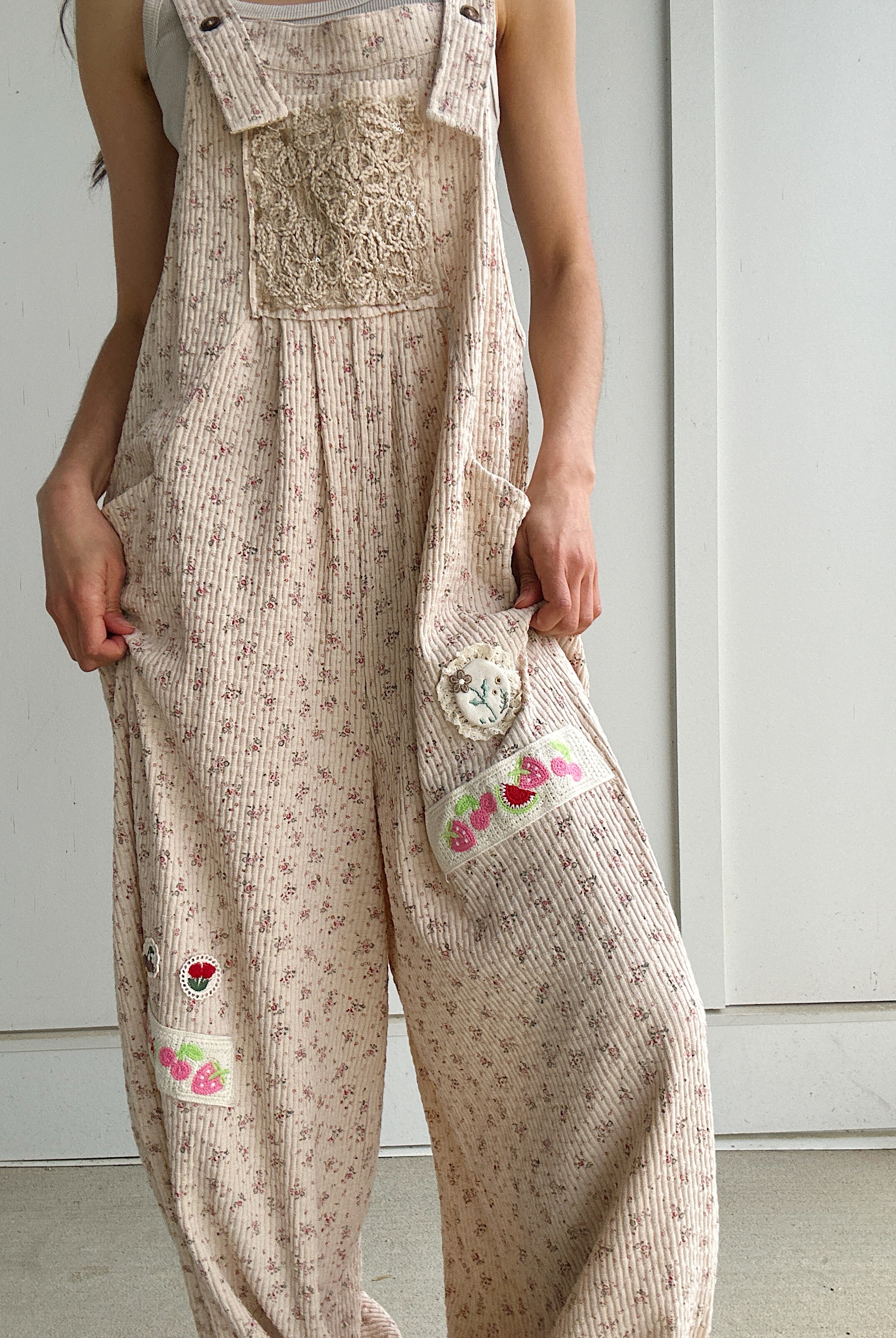 Person wearing beige floral overalls with decorative fruit and flower patches on a plain background