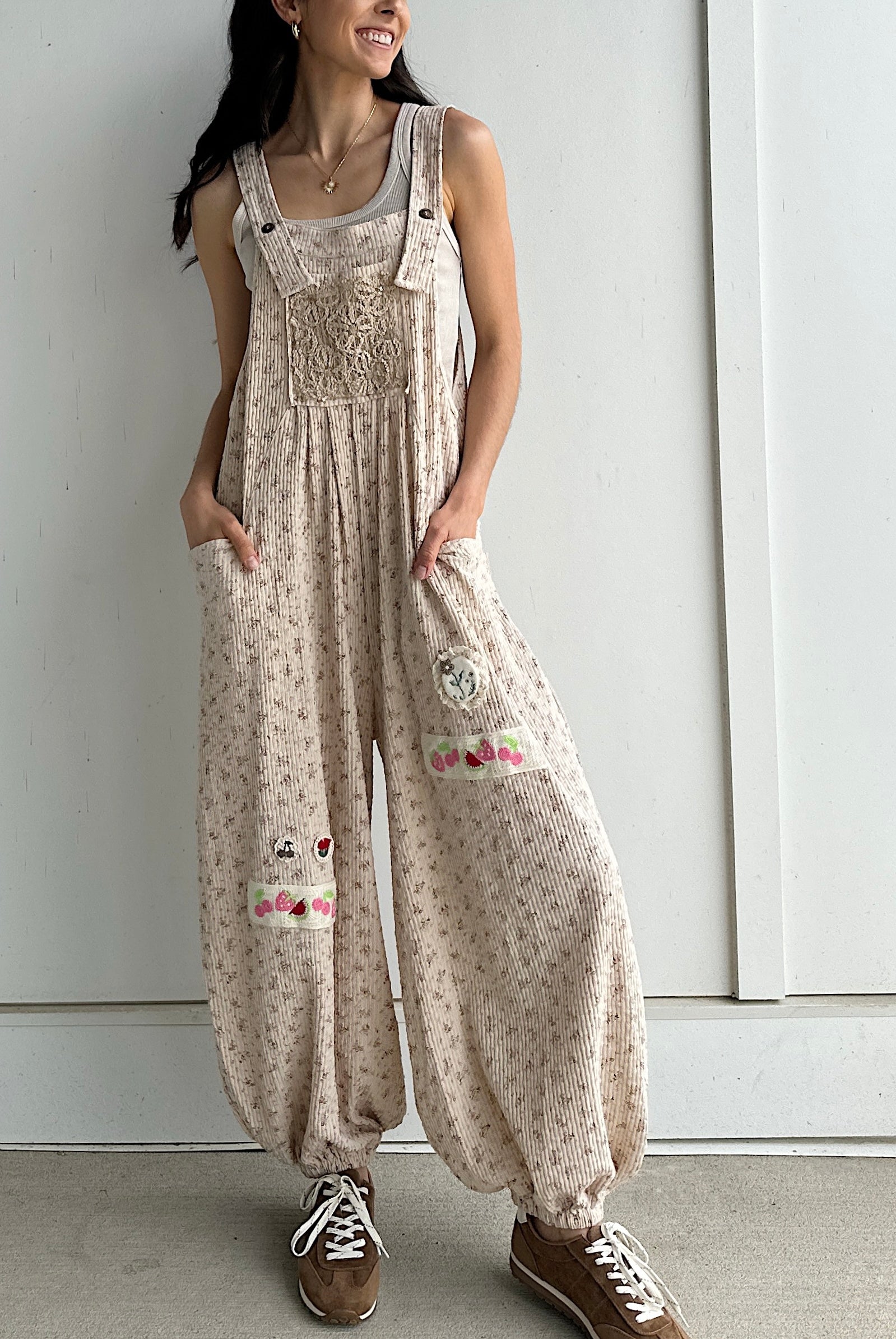 Person wearing a beige floral overalls with patches against a white wall.